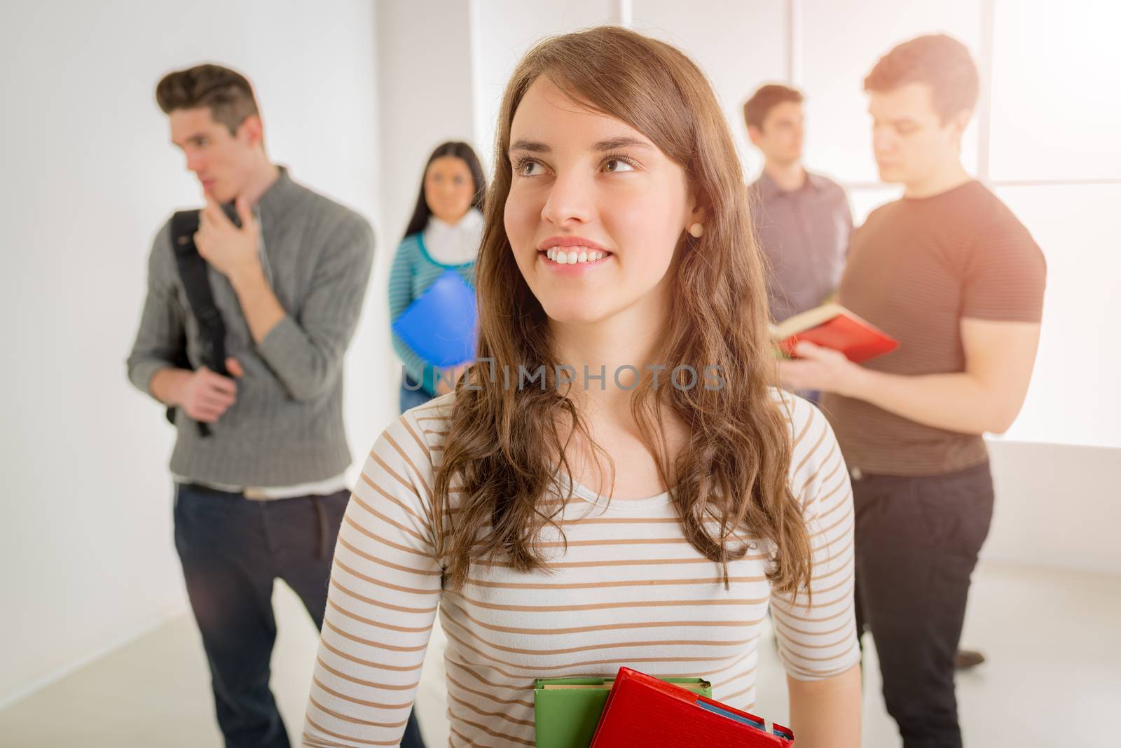 Beautiful pensive student girl with book standing in school hall. A happy group of his friends is behind him. 