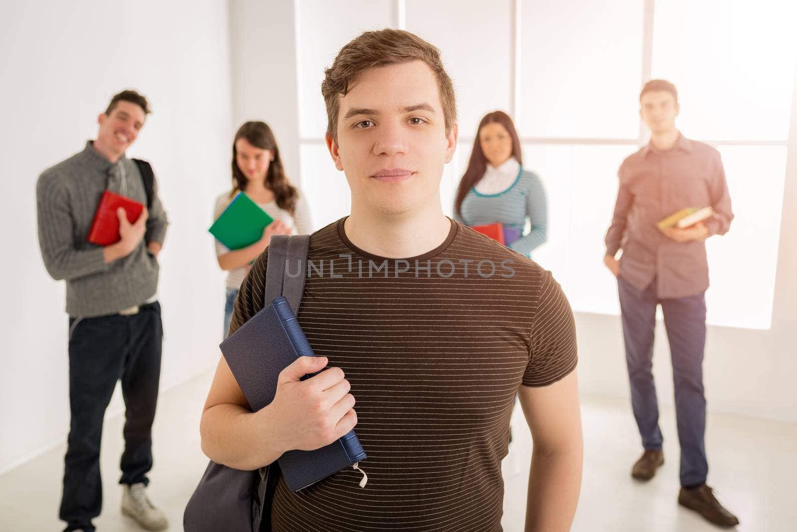 Beautiful smiling young man standing in school hall and looking at camera. A happy group of his friends is behind him. 