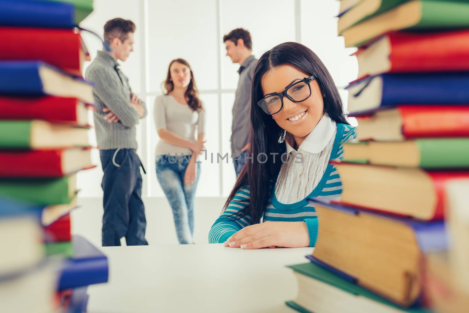Portrait of a beautiful smiling girl with glasses sitting behind the many books in the foreground. Looking at camera. A her friends standing  is behind her and talking.