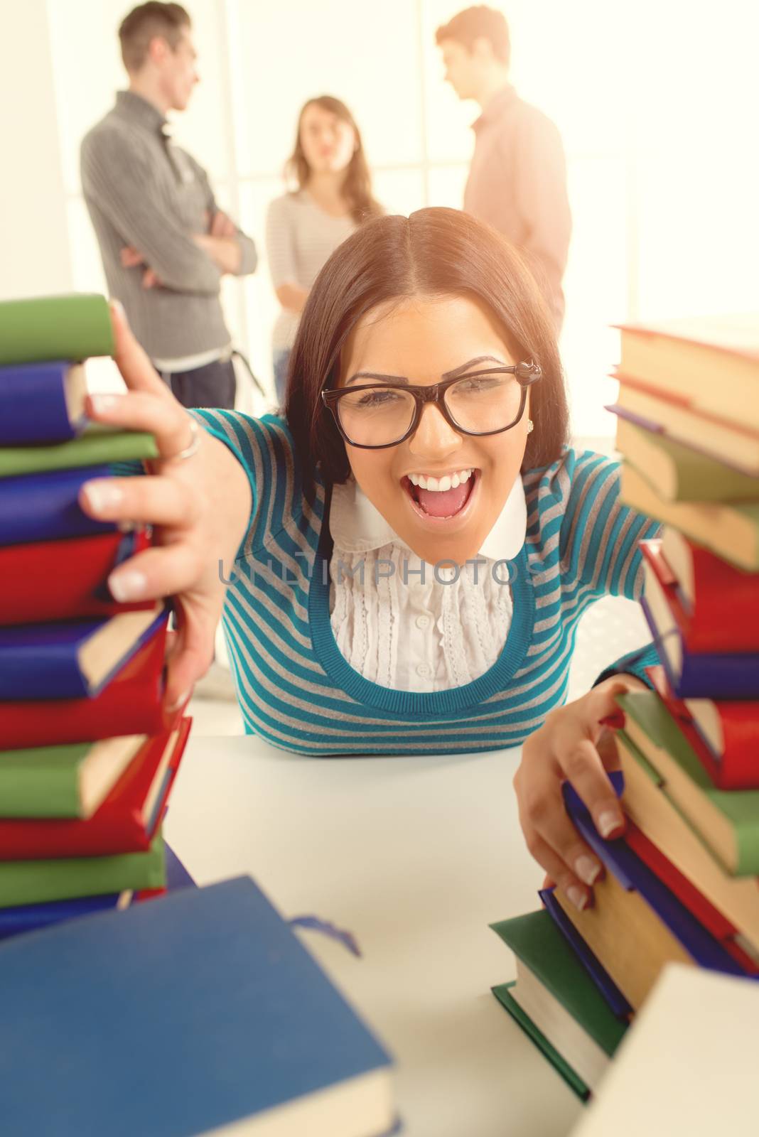 Portrait of a beautiful funny student girl sitting and shouting among the many books in the foreground. A her friends standing  is behind her and talking. Looking at camera.