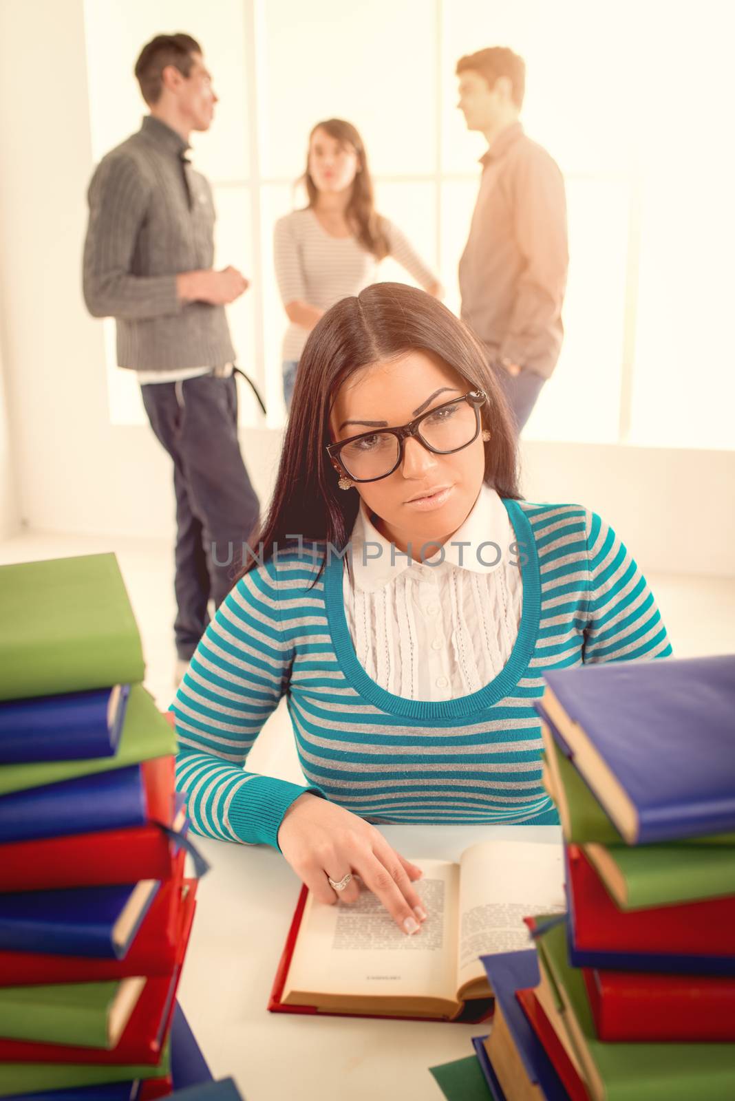 Portrait of a beautiful girl sitting and learning among the many books in the foreground. A her friends standing  is behind her and talking. Looking at camera.