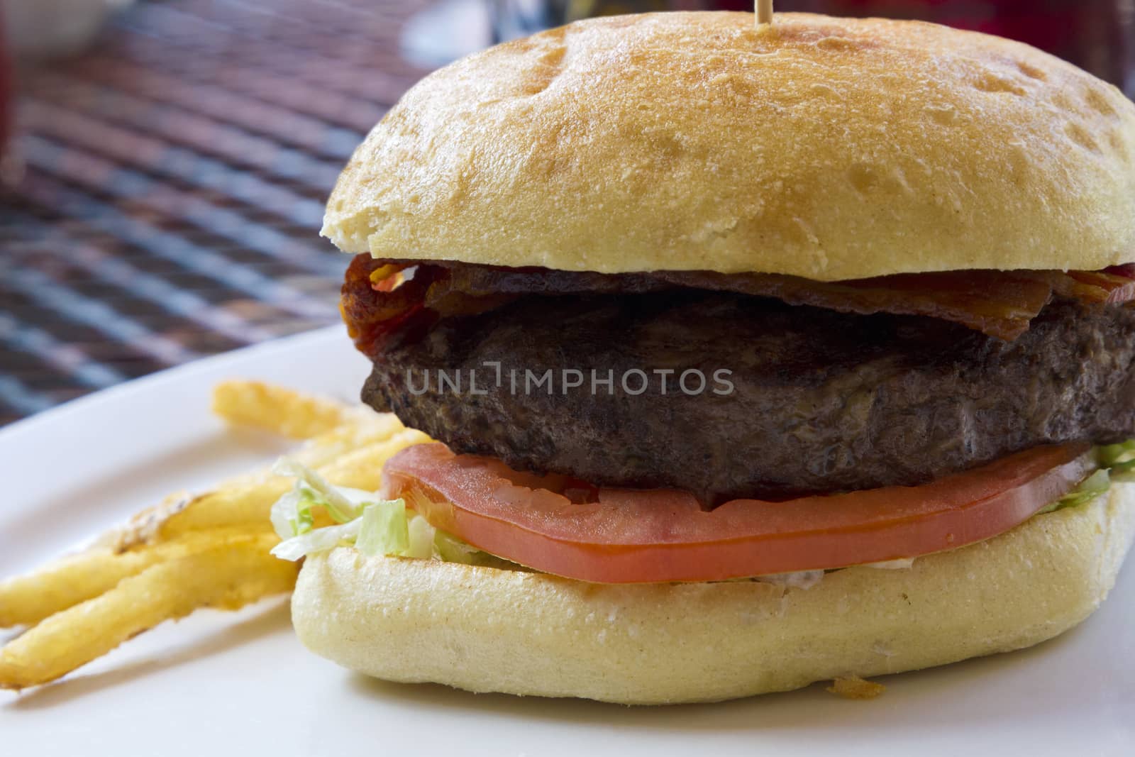 Juicy, grilled hamburger with cooked bacon slices, shredded lettuce, and a fresh tomato slice.  Meat on bread roll with fried potatoes on outdoor, wrought iron patio table in horizontal photograph. 