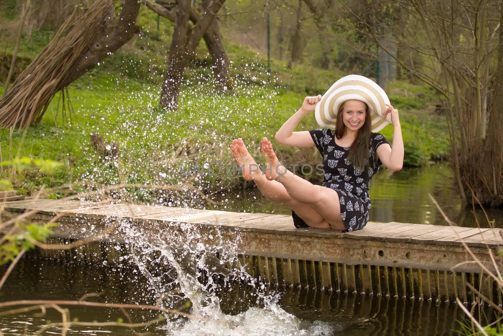 Cheerful fashionable woman sits on small bridge and splashing water by her legs in warm spring day.