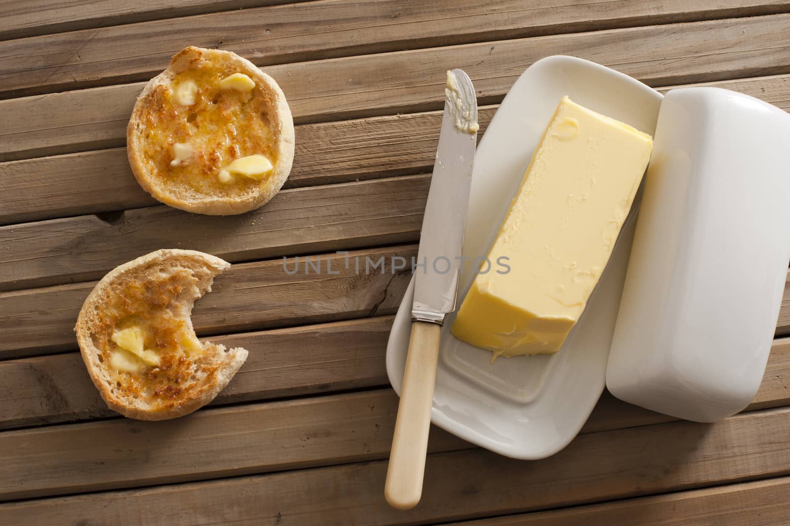 One yellow stick of butter on tray with knife besides two toasted english muffins on wood slat table
