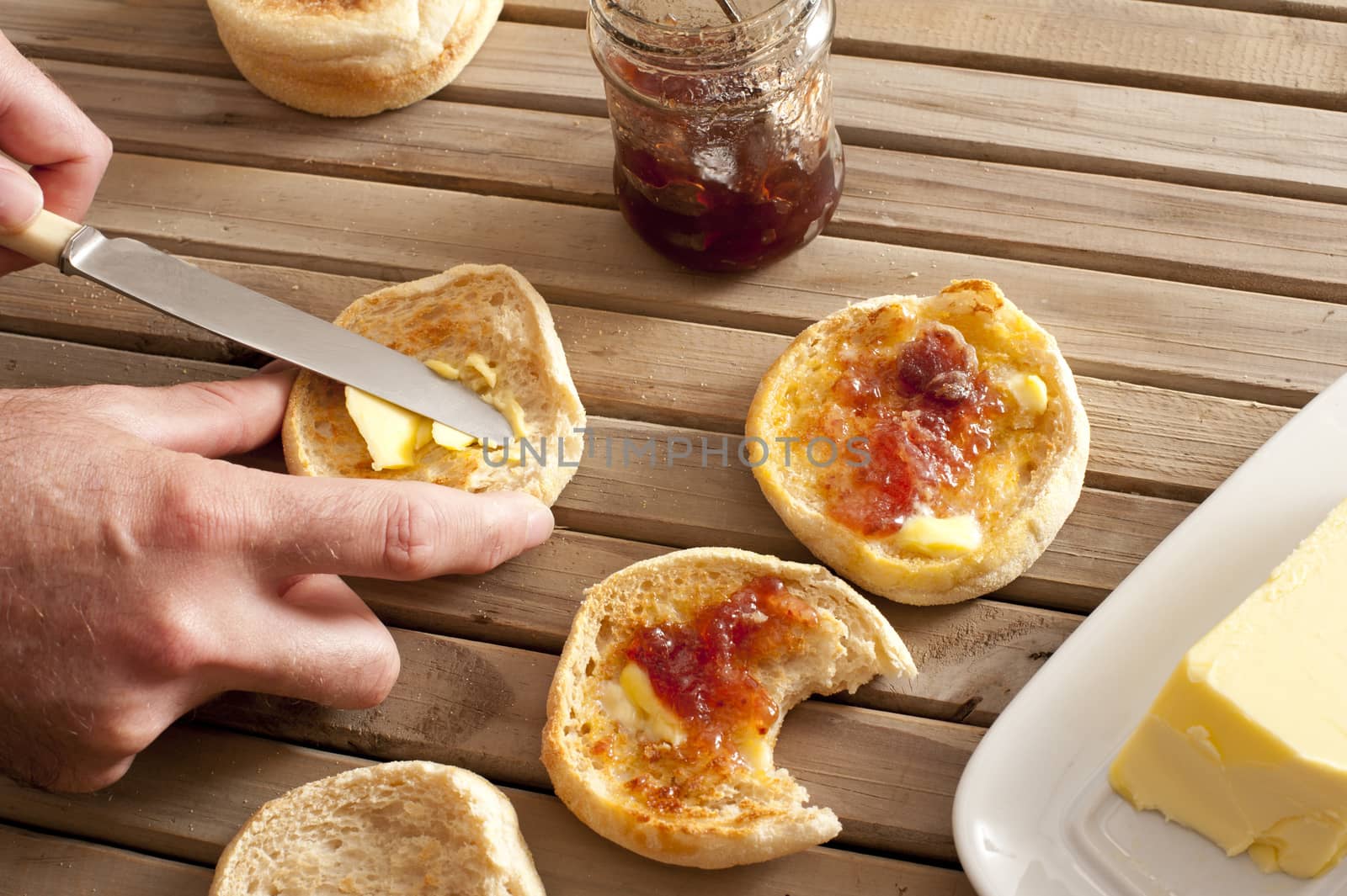 Pair of hands using knife to spread strawberry marmalade on toasted english muffins next to jelly jar and yellow stick of butter
