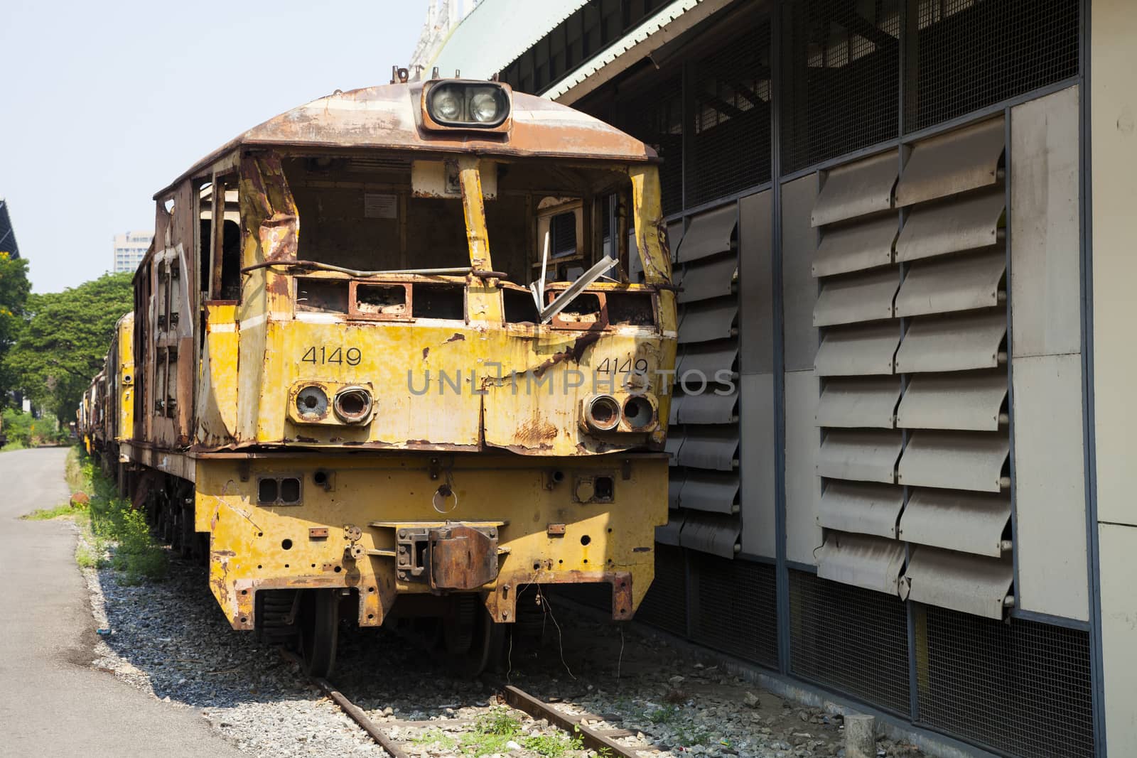 The ancient train has yellow or rust on the old railway near that has rocks and grass and behind has sky tree and buildings.