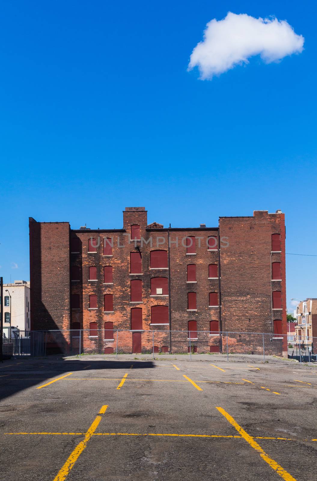 Yellow lines of an empty parking place and an abandoned building made of bricks. Bright blue sky with one single white cloud. Detroit, Michigan, USA.