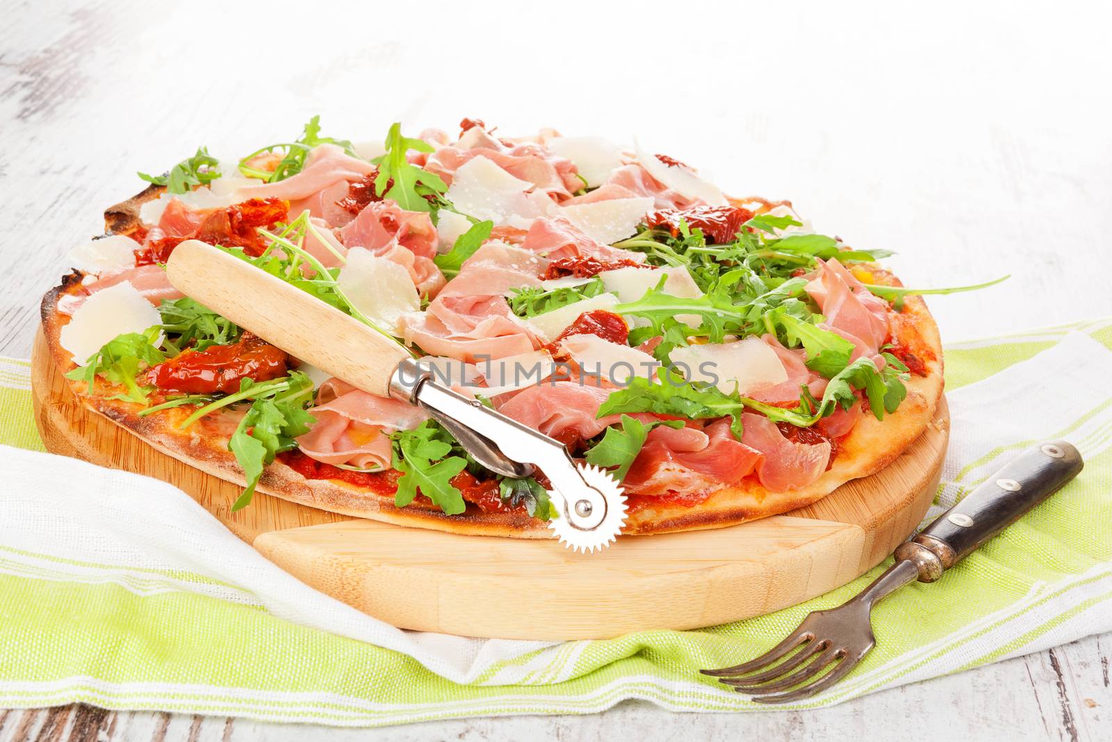 Culinary pizza with prosciutto, dry tomatoes and fresh herbs on wooden cutting board on wooden table. 