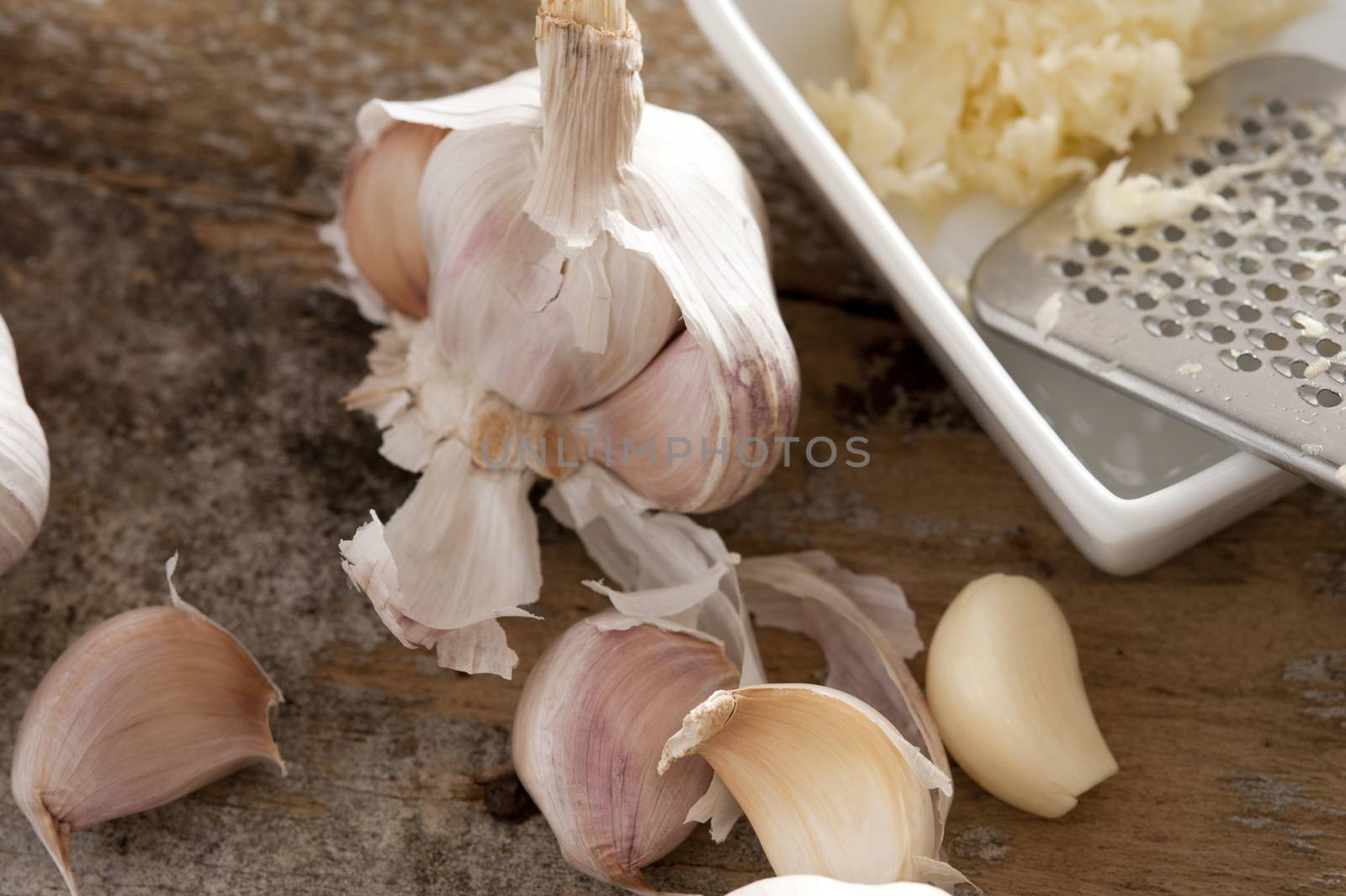 Preparing grated garlic for cooking with a fresh garlic bulb,, loose cloves both peeled and with skin on alongside a dish with a stainless steel grater