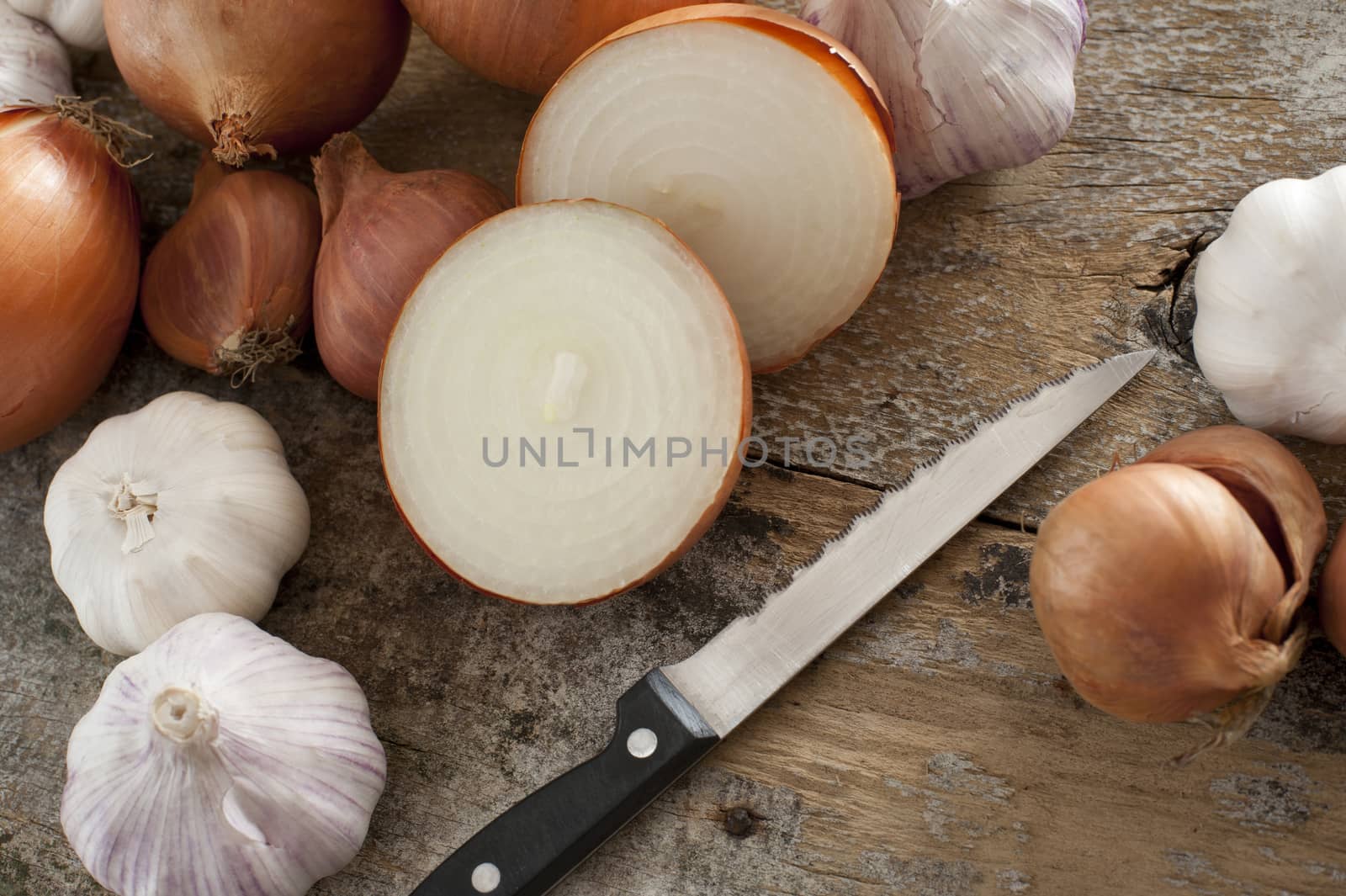 High Angle Still Life of Bounty of Onions and Garlic Bulbs Being Sliced and Prepared by Sharp Knife on Rustic Wooden Table