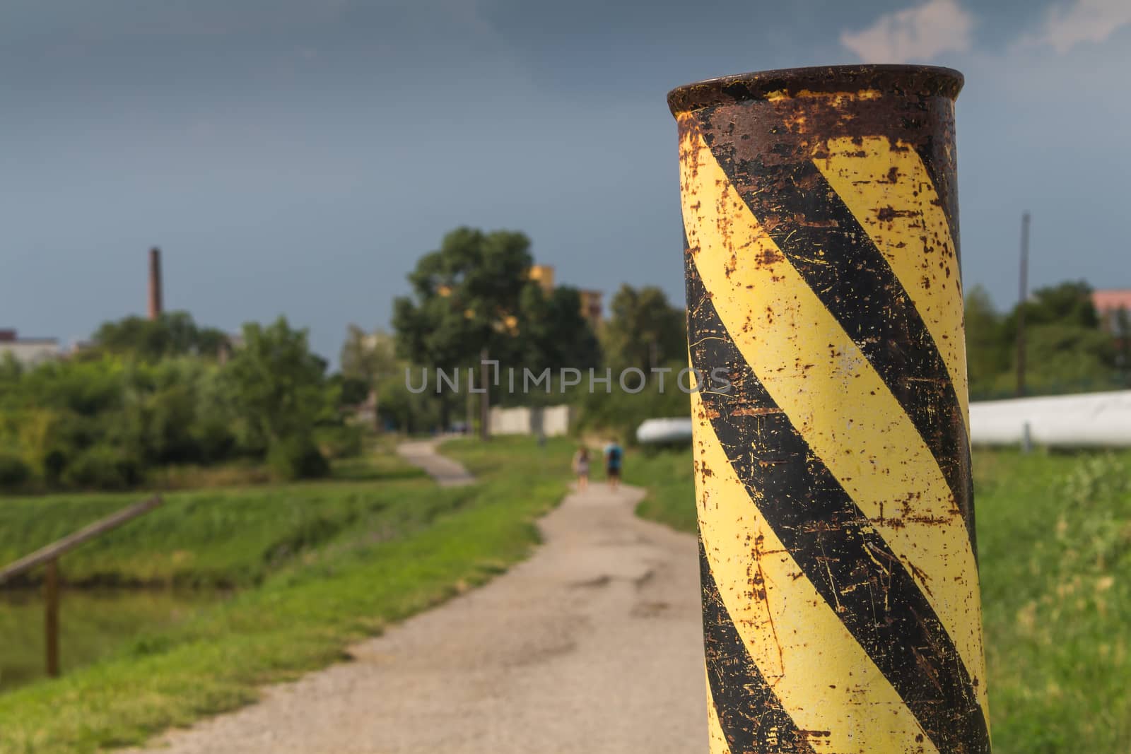 Detail of a rusted and column with black and yellow stripes, marking the beginning of a country road in a middle of a meadow with green grass. Stormy sky.