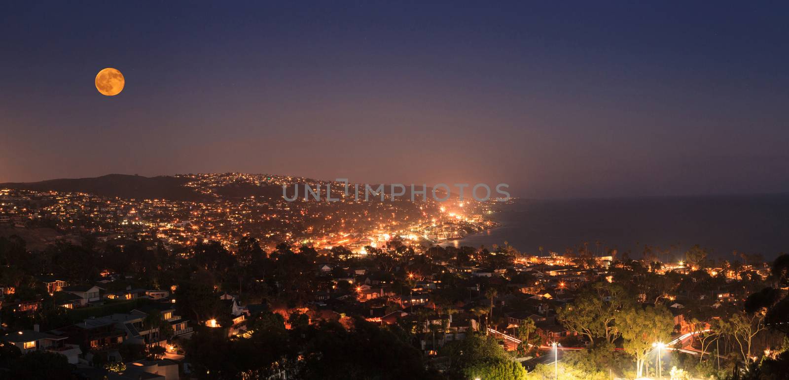 Moonrise of a full moon over the coastline of Laguna Beach, California, in summer on a clear night with city lights in the distance and the ocean at sunset.
