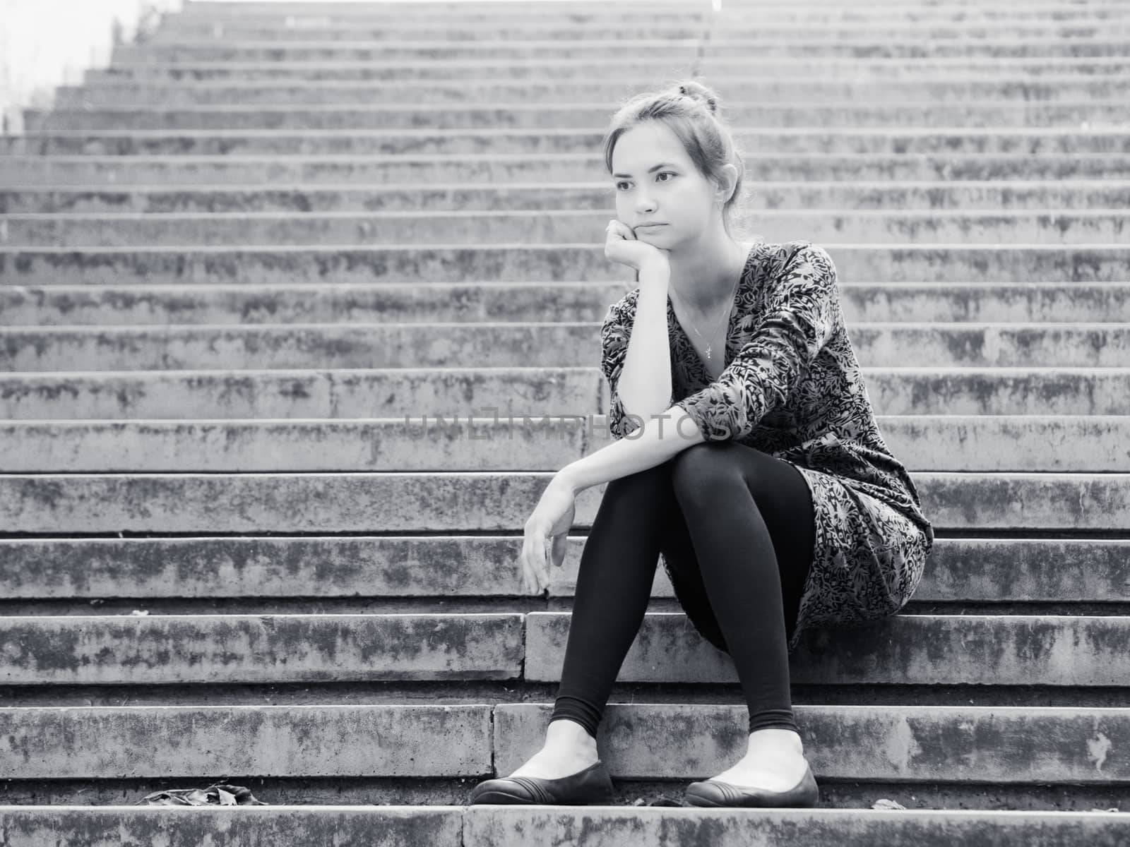 Sad lonely young woman sitting on steps. Portrait of serious girl with facial expression outdoors. Black and white image