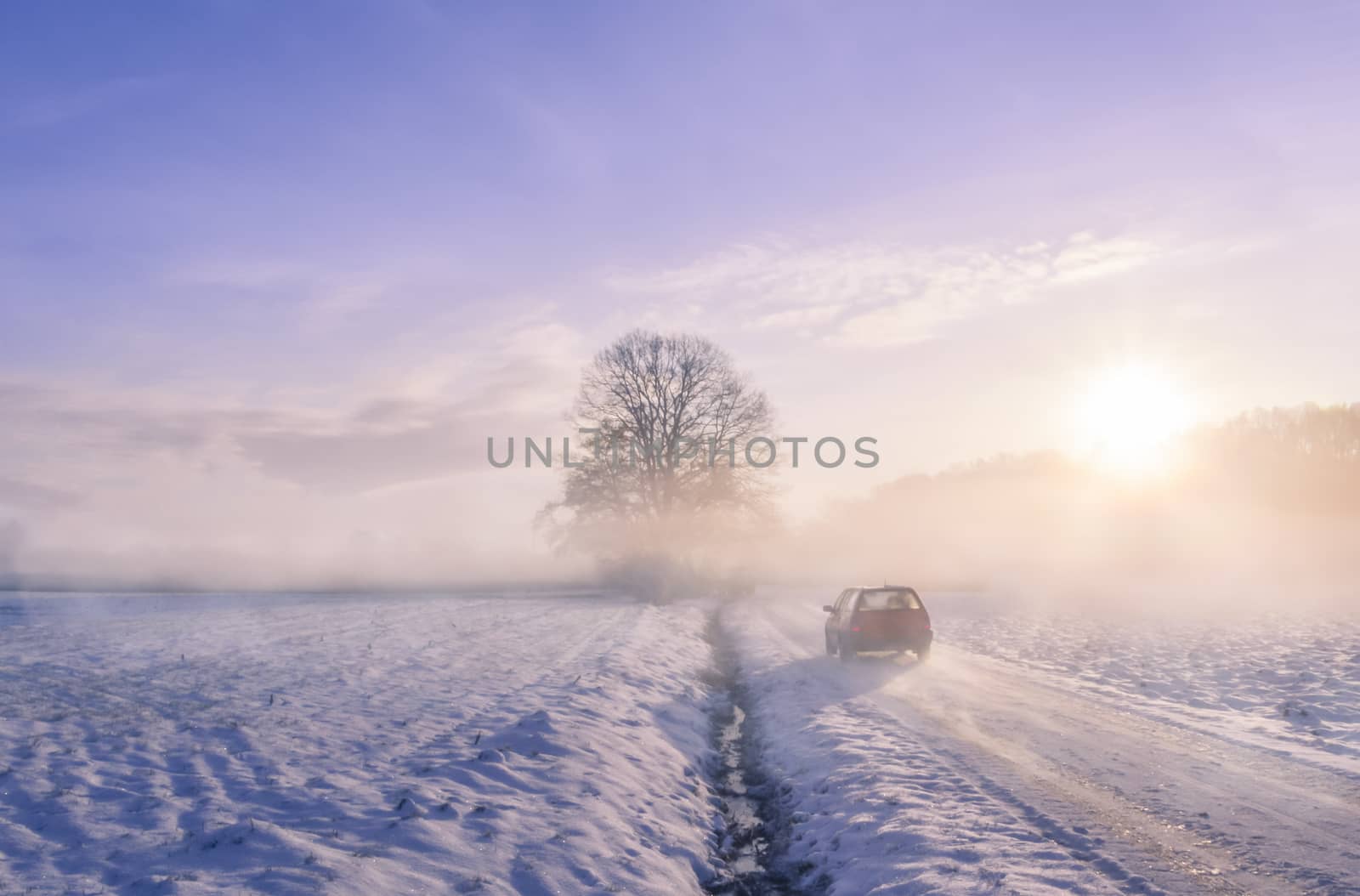 Winter image with the silhouette of a car driving on a snowy country road, through mist and the sunrise light.