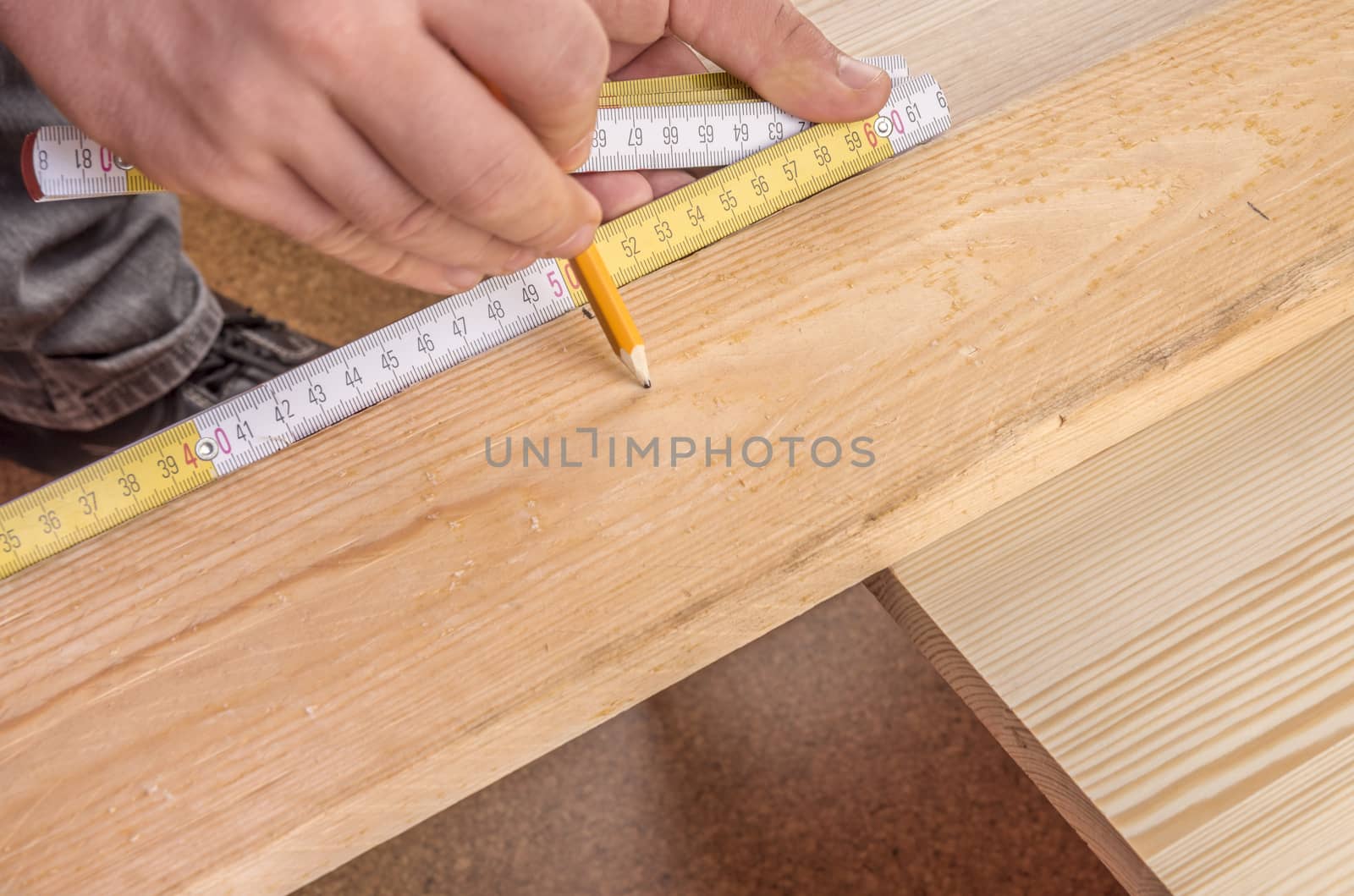 Close-up image with the hands of a man holding a measuring tape against a wooden board and makes a mark with a pencil.