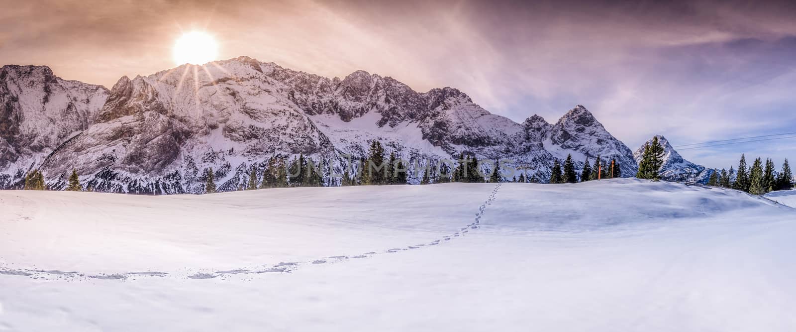 Winter landscape with Austrian alps peaks, a single trail of footsteps on snow, under a dramatic and colorful sky.