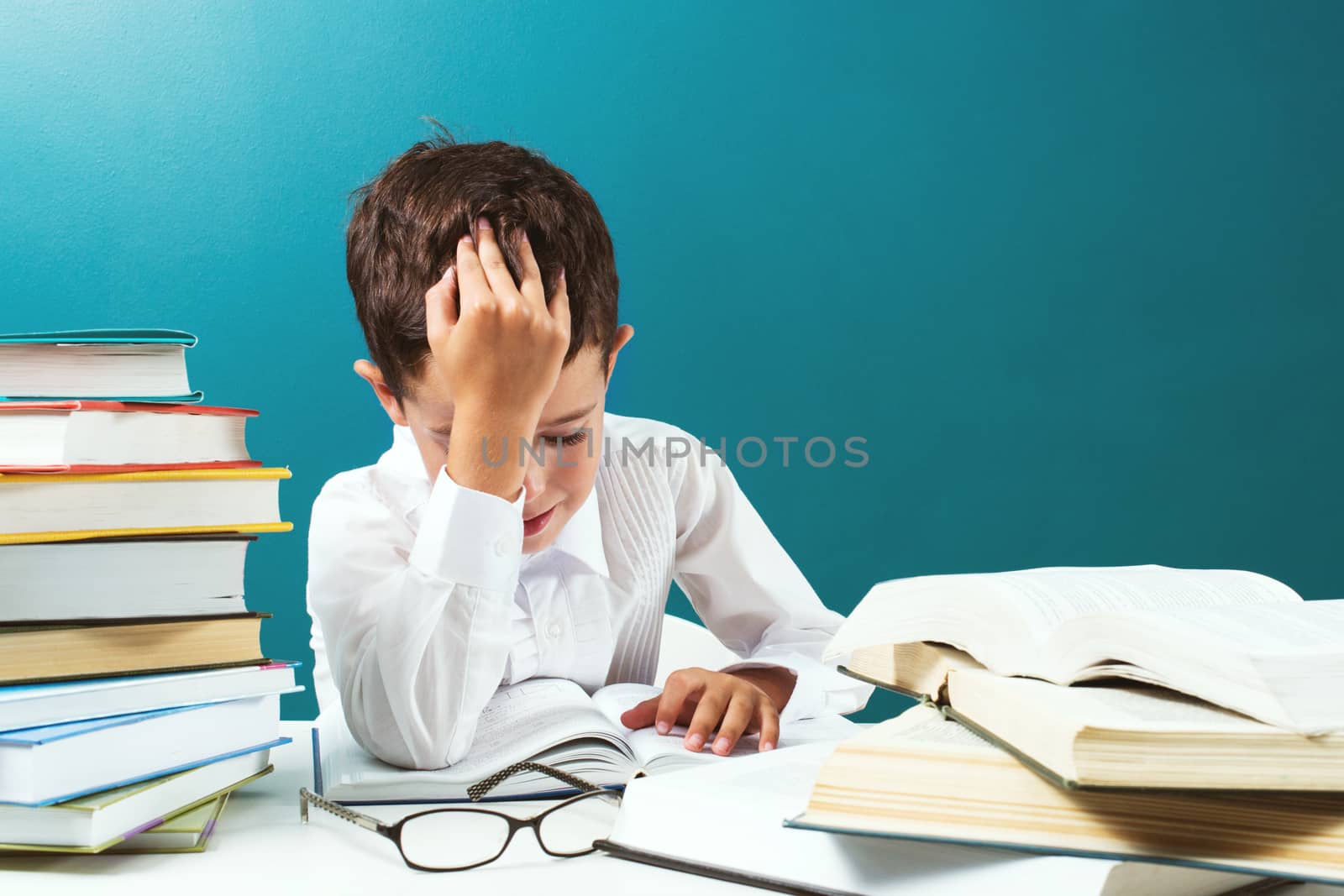 Сute boy reading difficult  book at the table, blue background