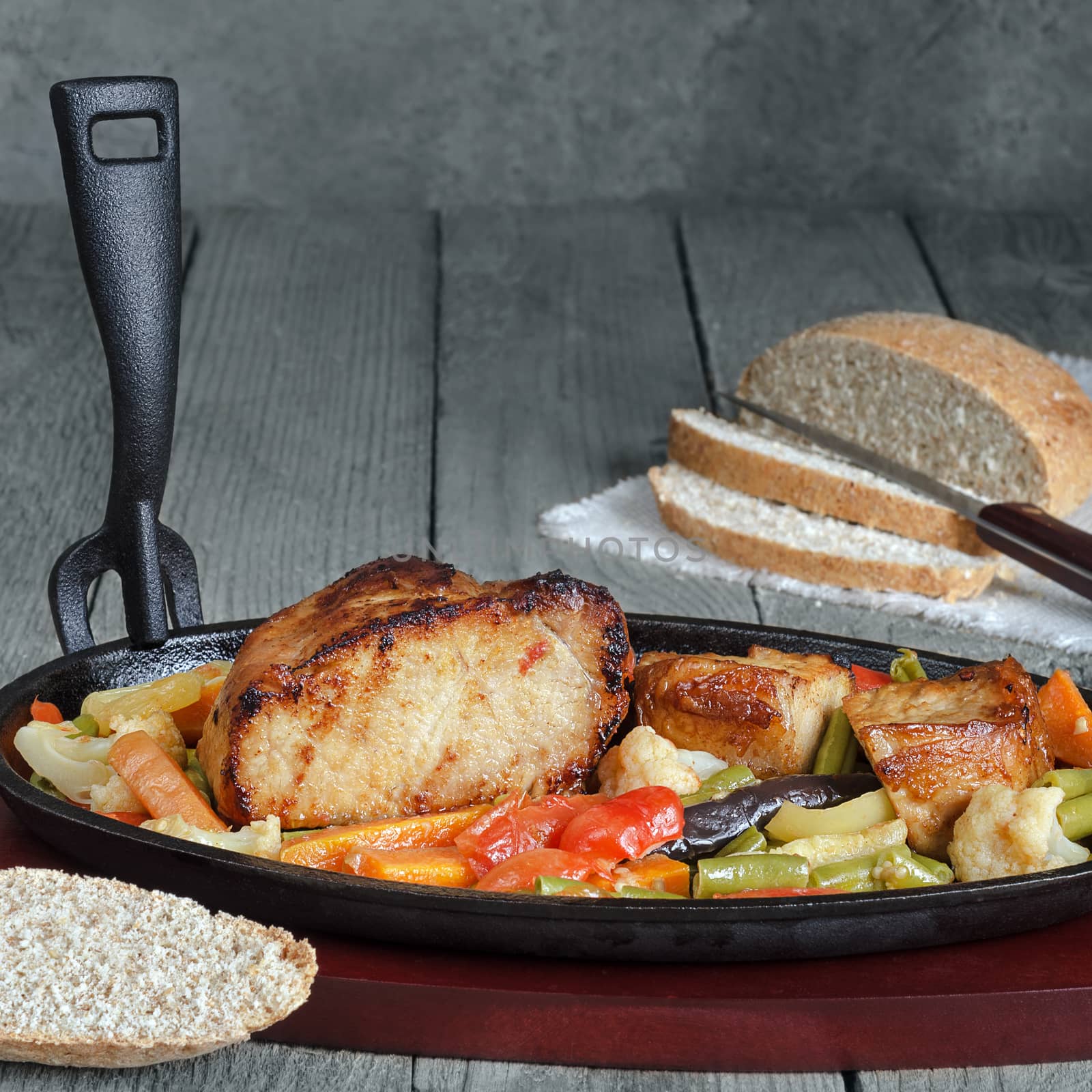 Fried pork with vegetables in a cast iron skillet and a wooden stand. Blue gray wood background, rustic style.