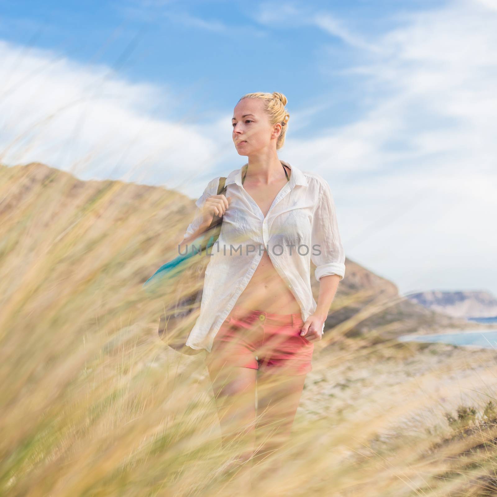 Relaxed woman on vacations in white loose shirt carrying beach bag and towel, enjoying beautiful coastline view of white sandy lagoon at Balos beach, Greece.