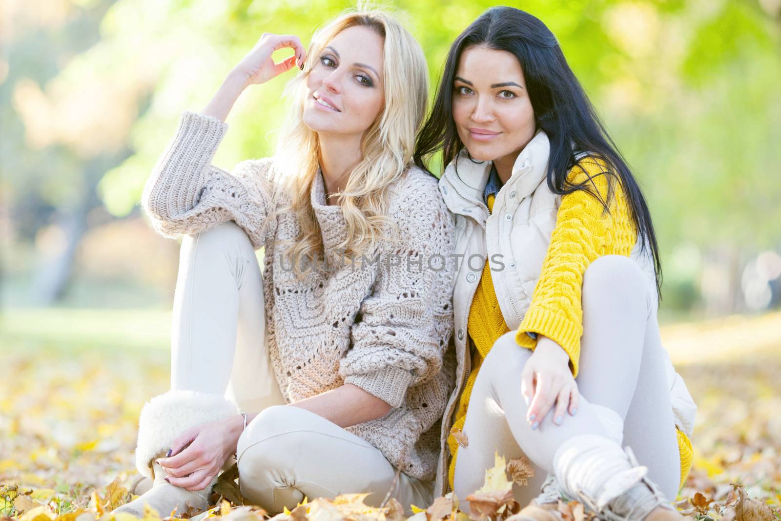 Two cheerful women sitting on dry leaves in autumn park at sunny day