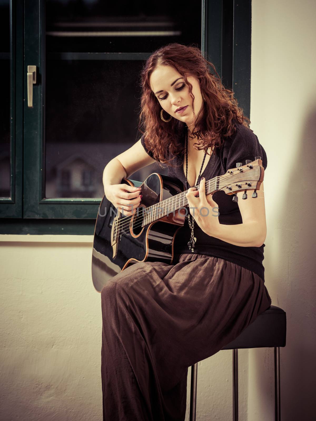 Photo of a beautiful brunette female playing an acoustic guitar by the window.
