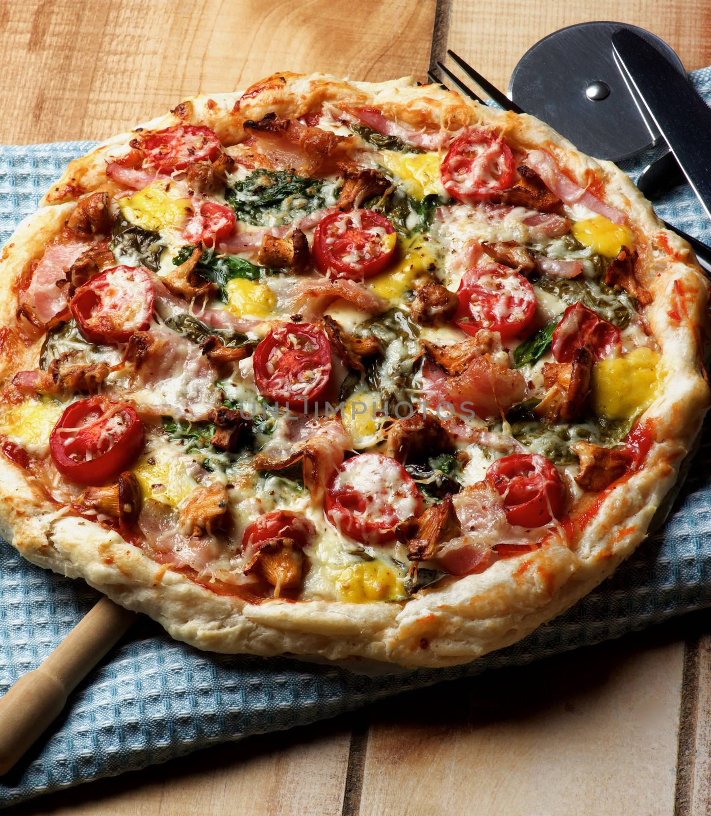 Delicious Mushrooms Pizza with Forest Chanterelles, Bacon, Tomatoes, Basil and Cheese on Blue Napkin with Pizza Cutter closeup on Wooden background