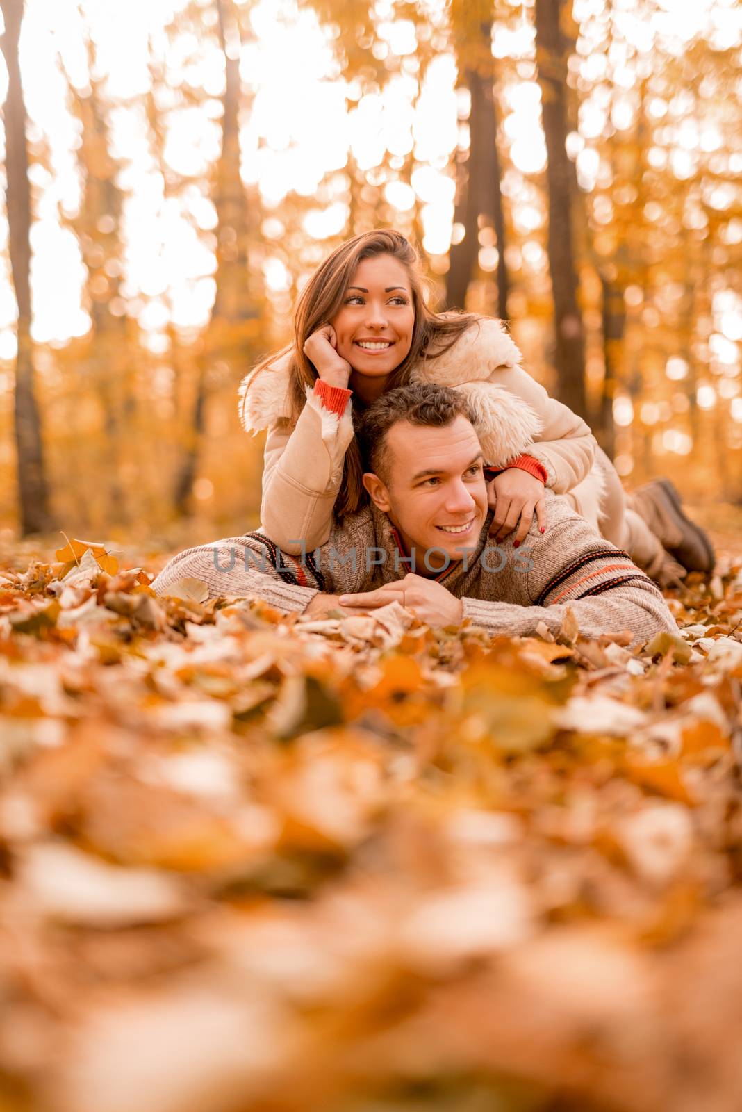 Beautiful smiling couple enjoying in sunny forest in autumn colors. They are lying on the falls leaves and looking away.