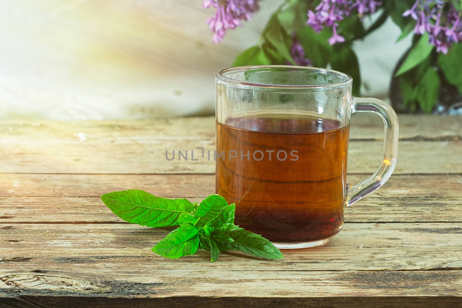 Tea in glass mug and fresh mint leaves on a rough wooden background. In the background - blurred flowers and leaves of lilac. Selective focus color image with copyspace