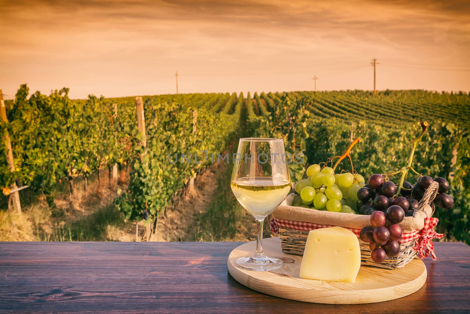 Glass of white wine with grapes in a basket and cheese in front of a vineyard at sunset
