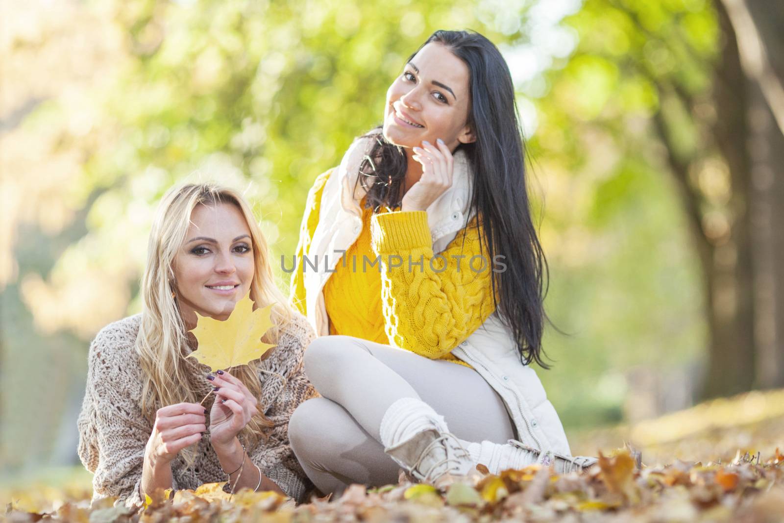 Two cheerful women sitting on dry leaves in autumn park at sunny day