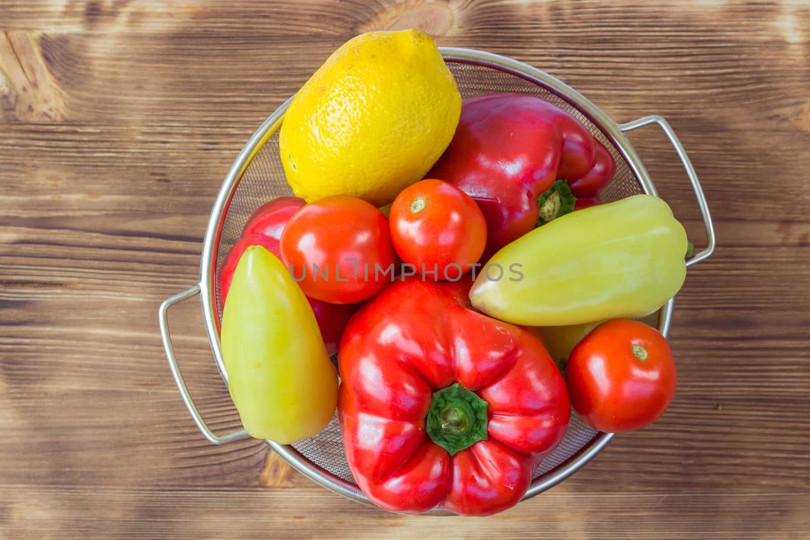 Vegetable still life of red and green peppers, tomatoes and lemon in sieve bowl on wooden board background