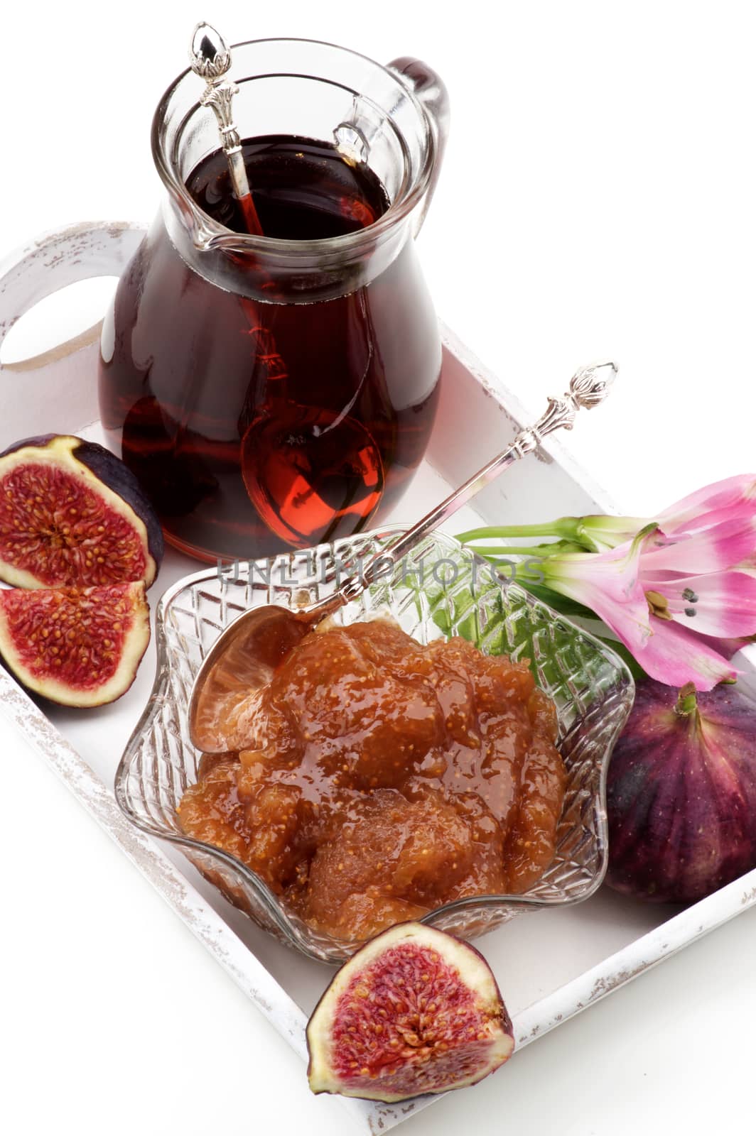Arrangement of Delicious Homemade Fig Jam in Glass Kremanka with Fresh Fig Fruits, Flowers and Jag of Fruit Drink in White Wooden Tray closeup on White background