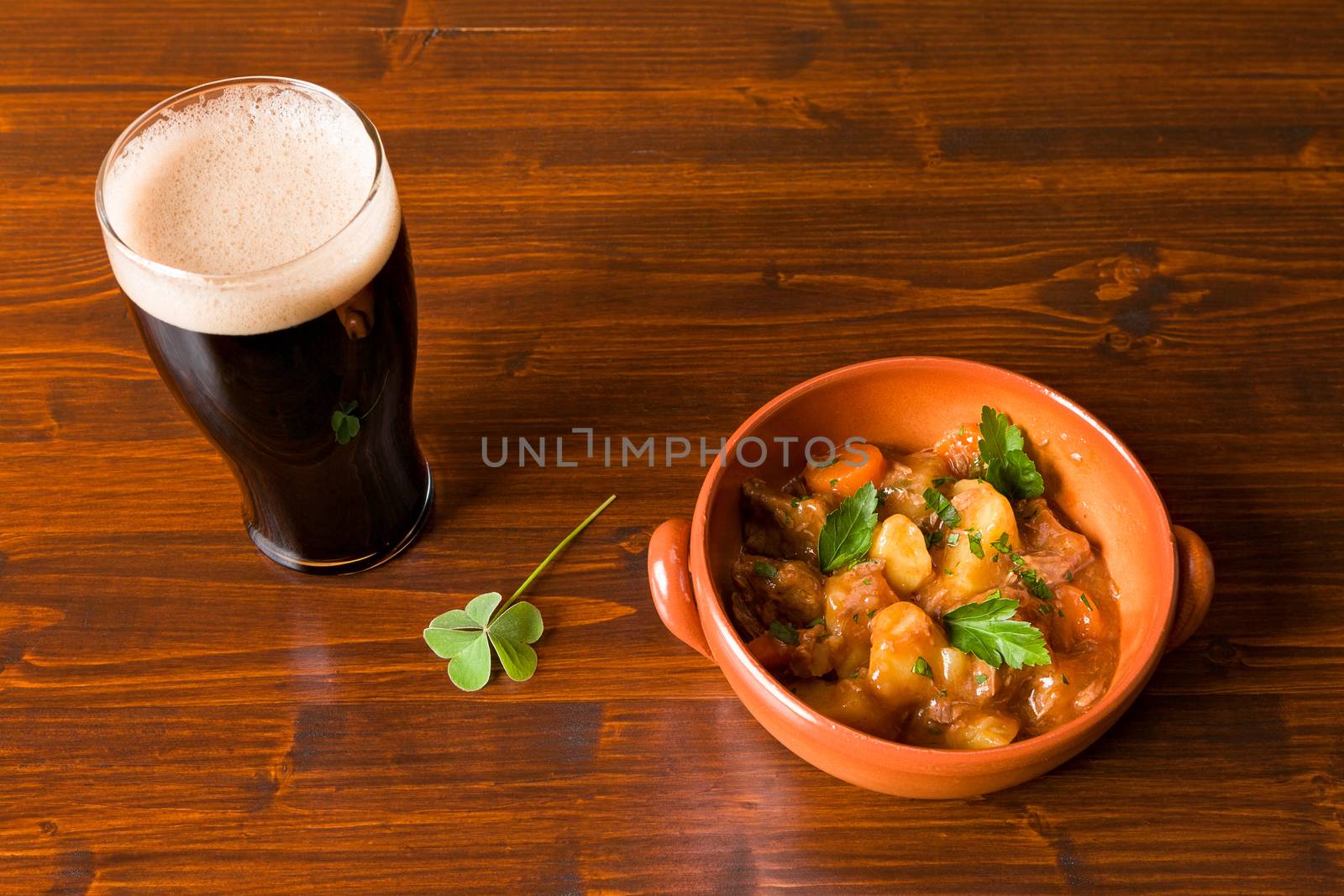 Traditional Irish Stew on a brown bowl with a pint of stout beer and a shamrock on a table.