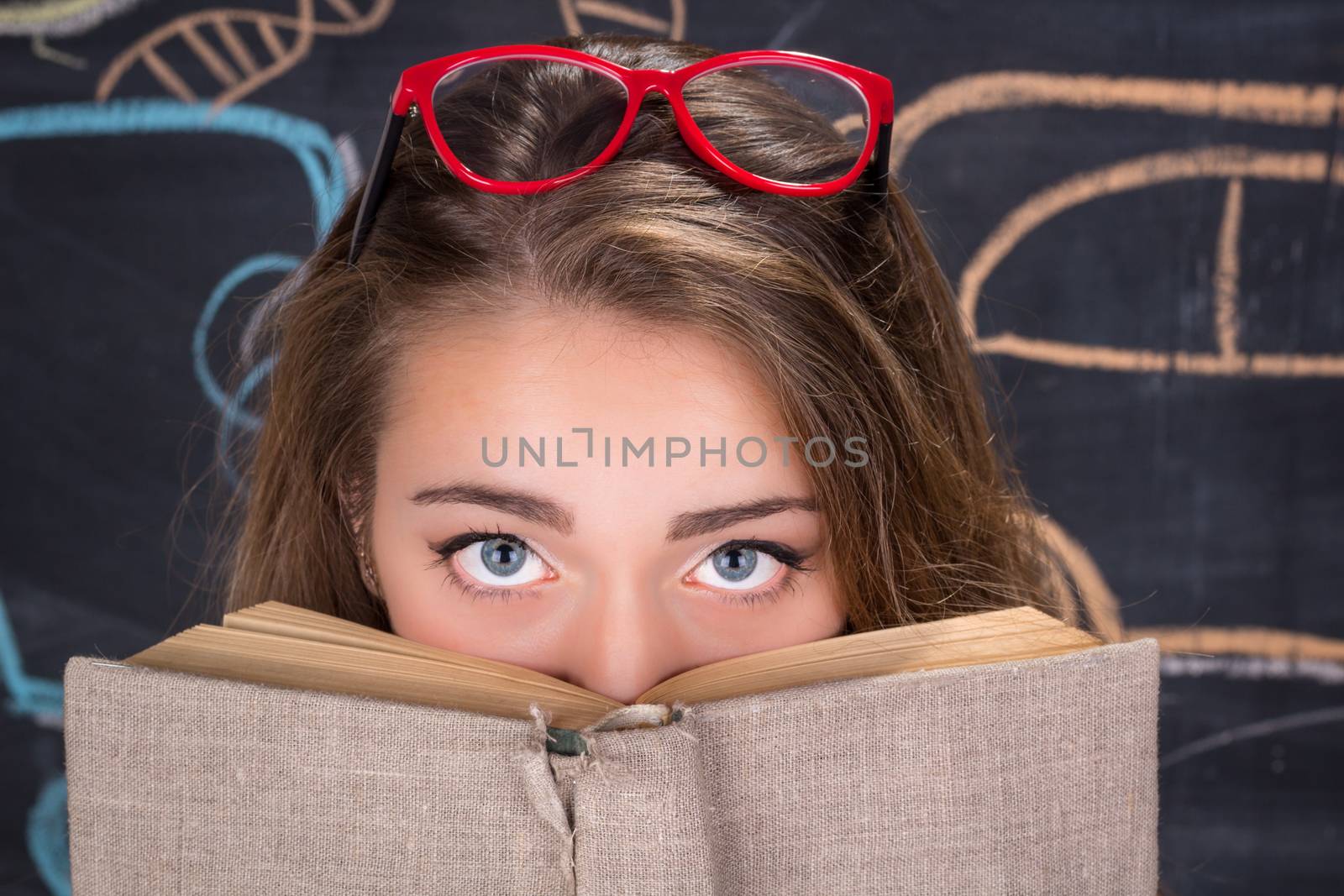 Perplexed  young student girl in red dress and red glasses reading a book hides behind it