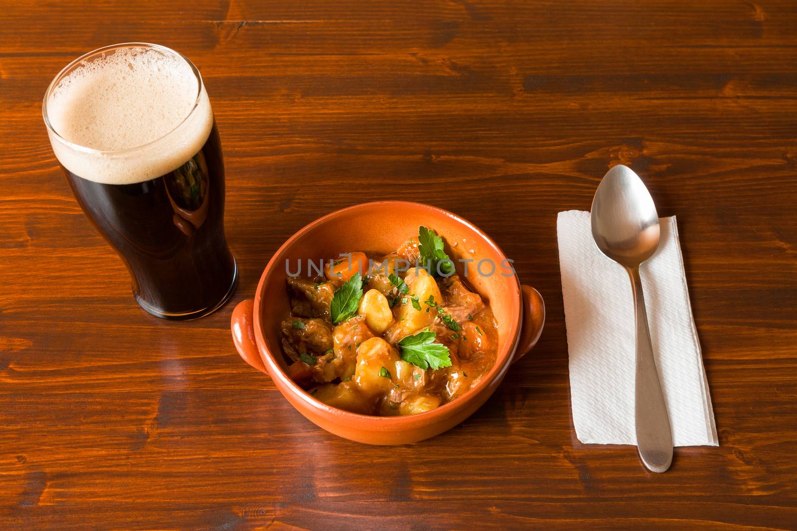 Traditional Irish Stew on a brown bowl with a pint of stout beer and a spoon over a napkin on a table.