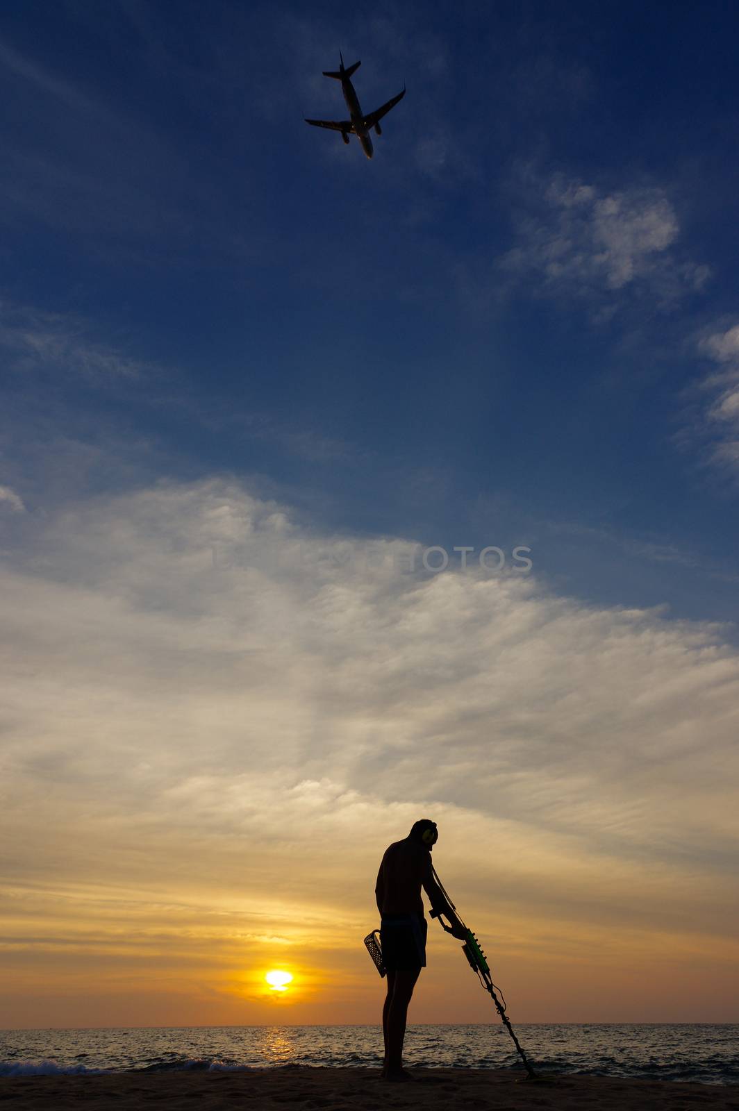 Treasure hunter with Metal detector on sunset  the beach  a plan in the sky by evolutionnow