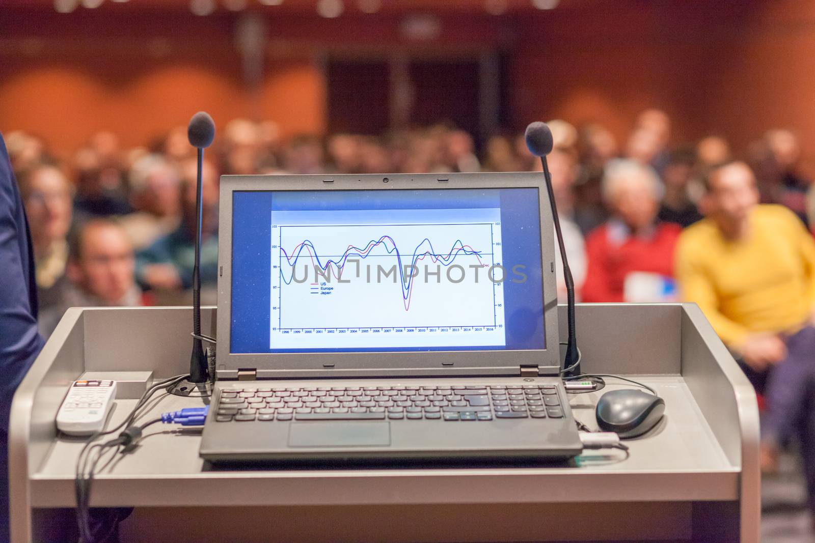 Computer and microphone on a podium desk at business event. Blured audience at the conference hall in background. Business and Entrepreneurship concept. Focus on computer.