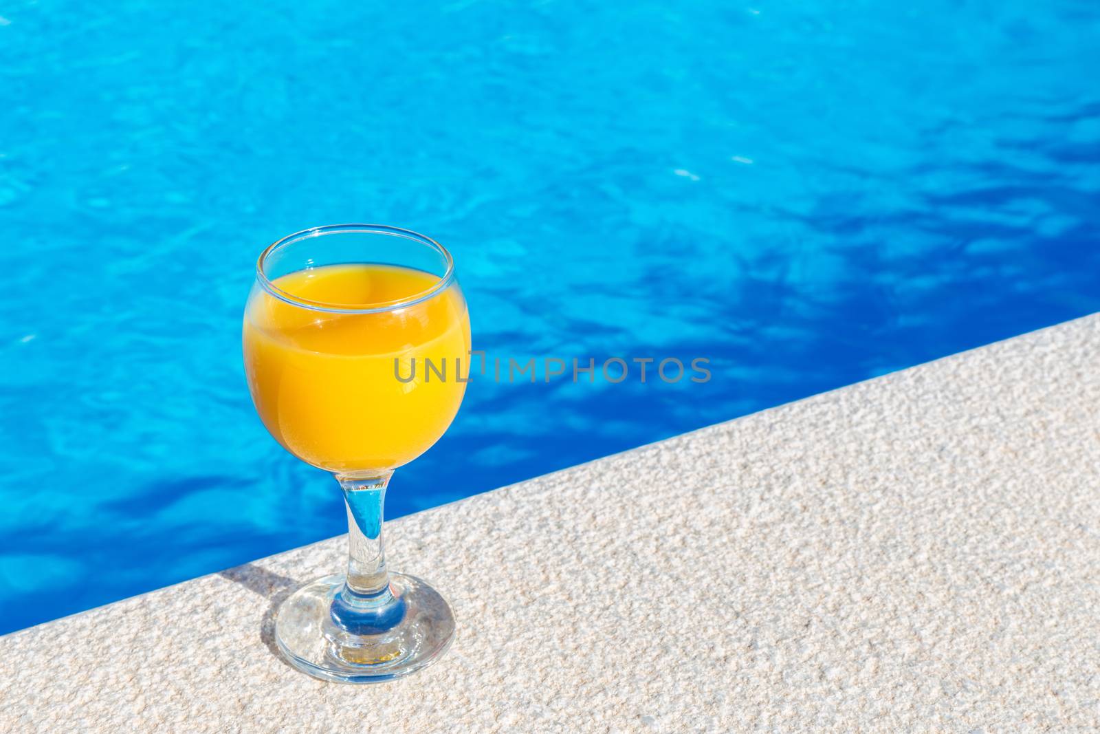 Glass filled with orange juice standing on edge of blue swimming pool