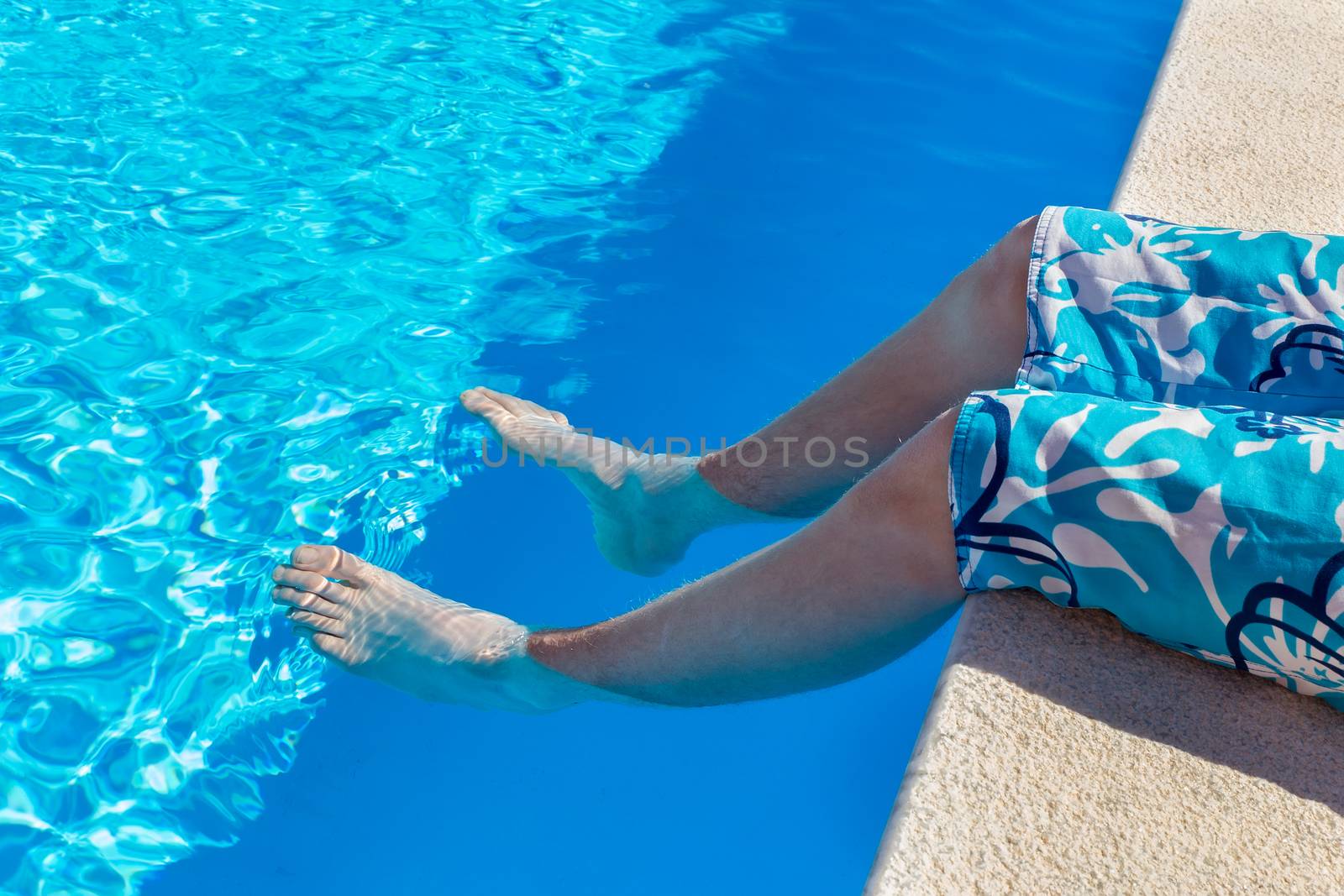 Teenage boy holding bare legs in blue swimming pool by BenSchonewille