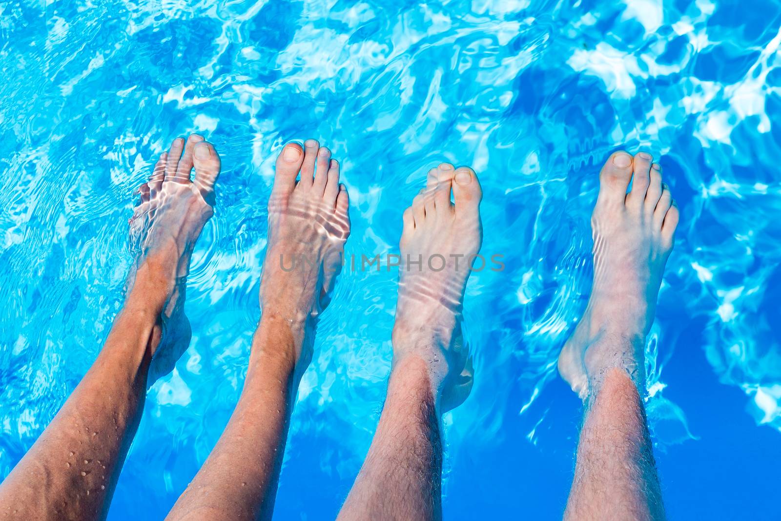 Four bare teenager legs with four feet in water of blue swimming pool