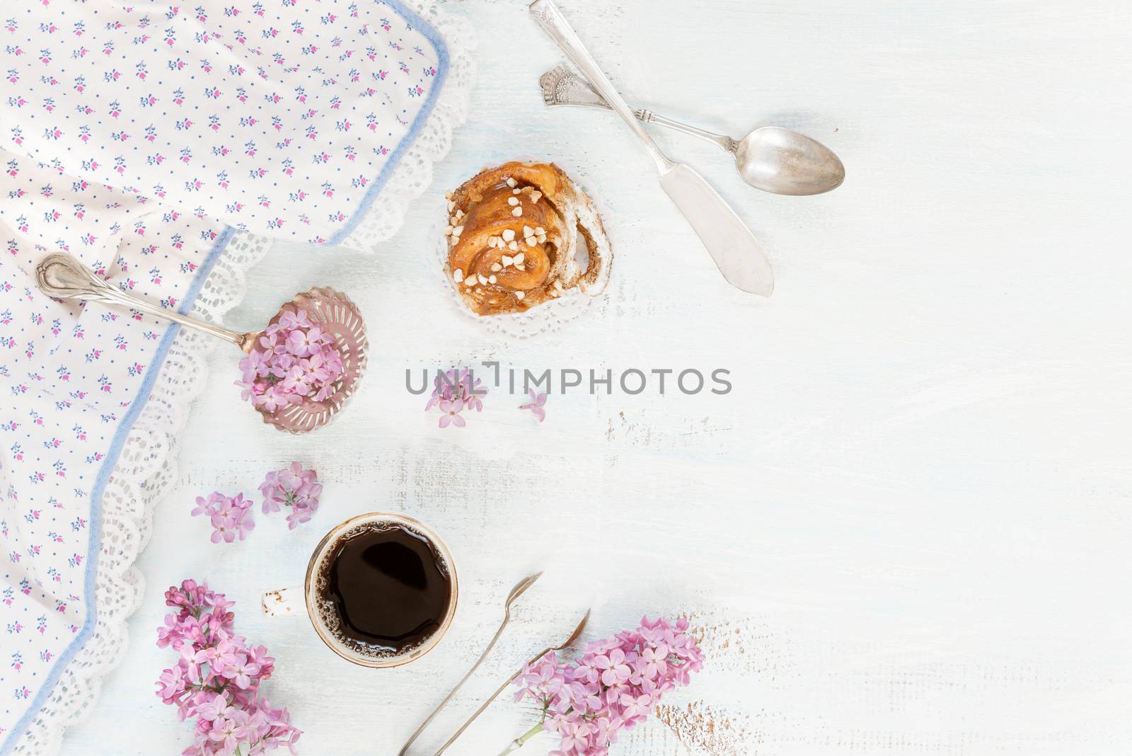 Breakfast consisting of a cup of black coffee and cinnamon bun, surrounded by lacy napkin and flowers of lilac on a light background; flat lay, top view, overhead view