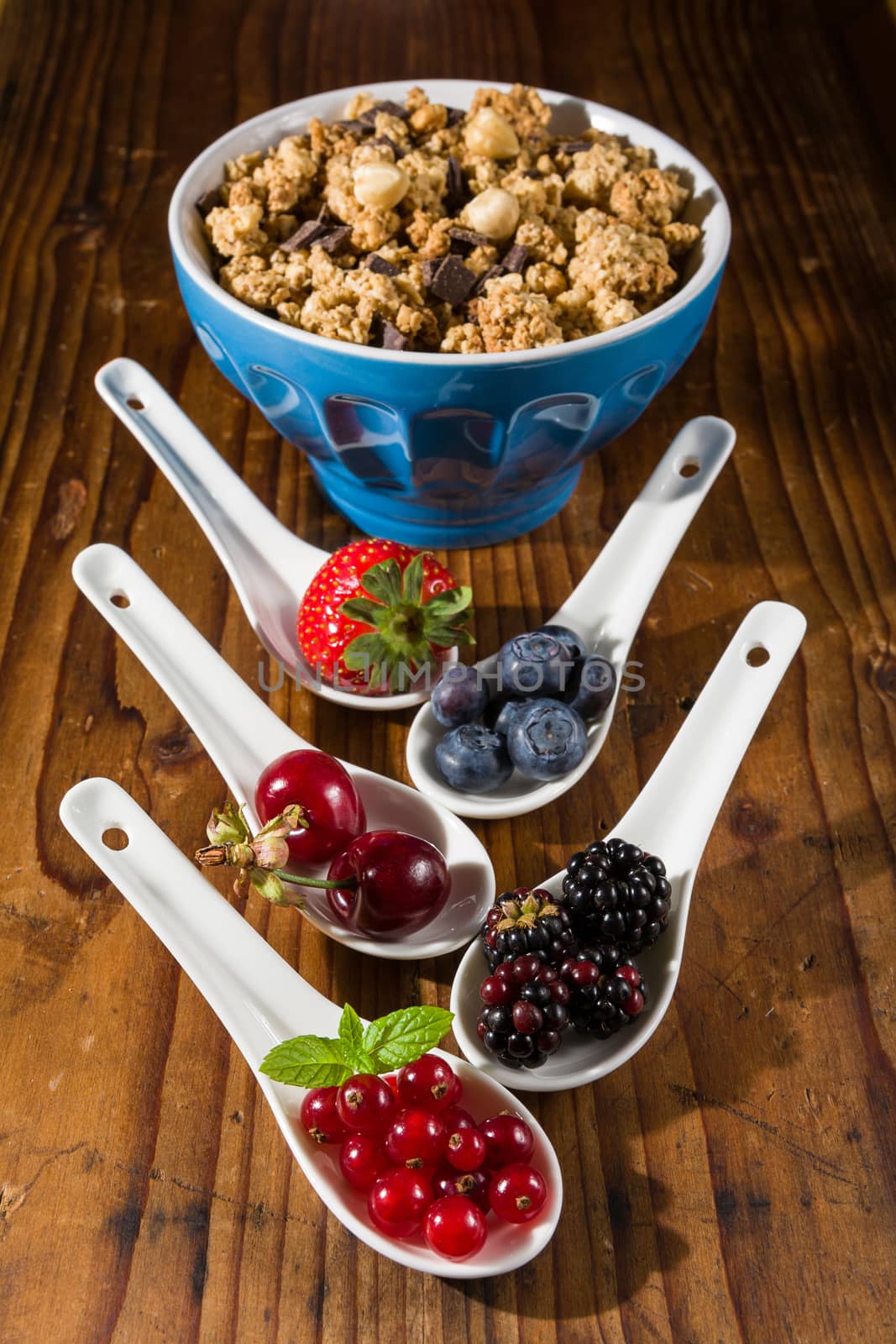 Collection of wild berries on white spoons and a cup of cereals over a wooden background