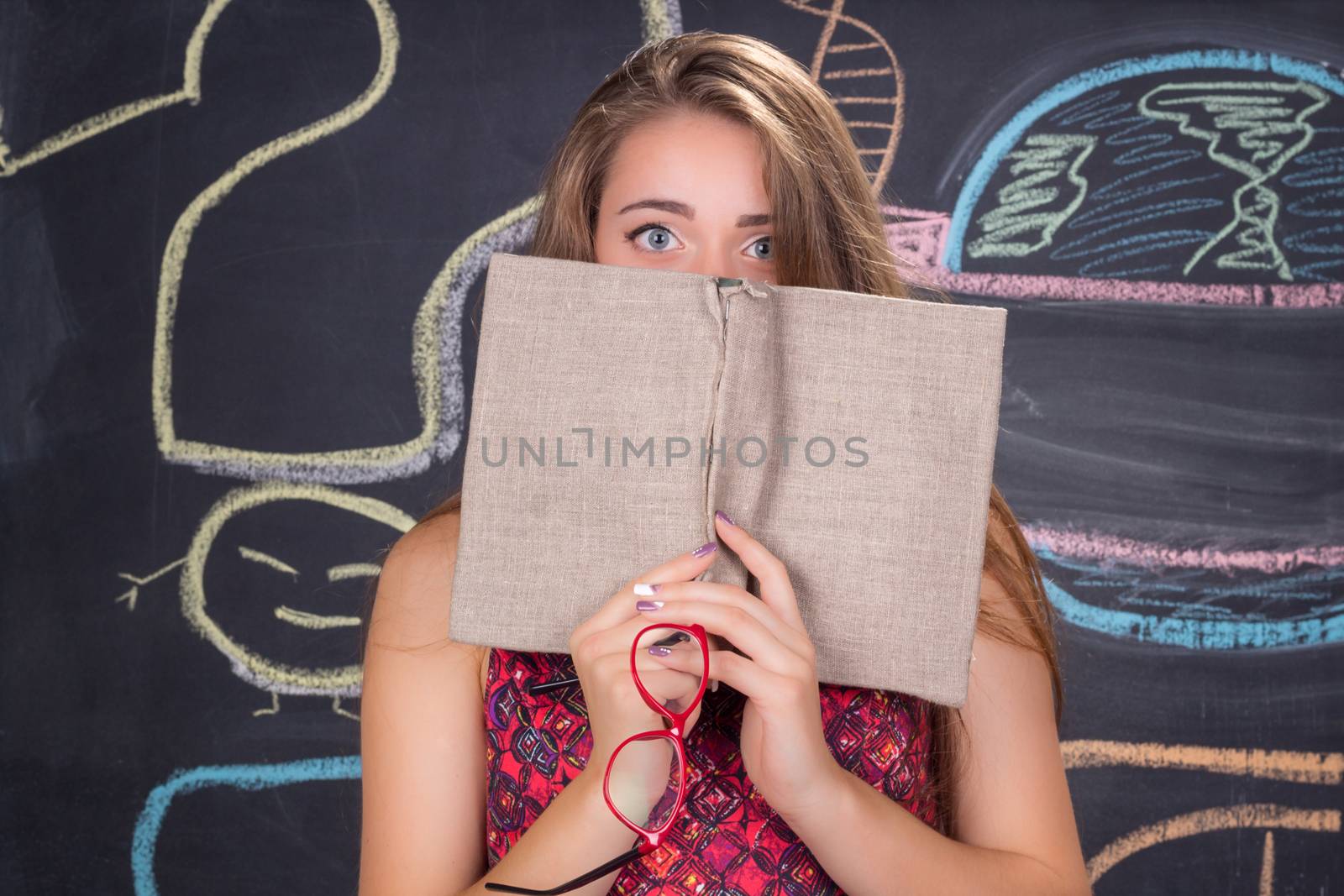 Perplexed  young student girl in red dress and red glasses reading a book hides behind it