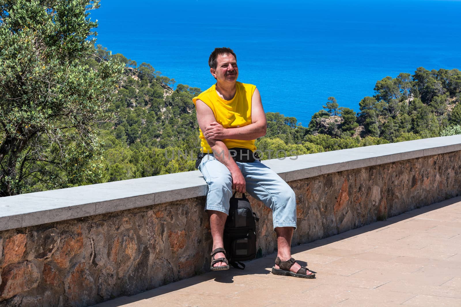 A man sitting in the sunshine on a wall in the background pine forest and the blue sea. Shot in Mallorca, Spain.