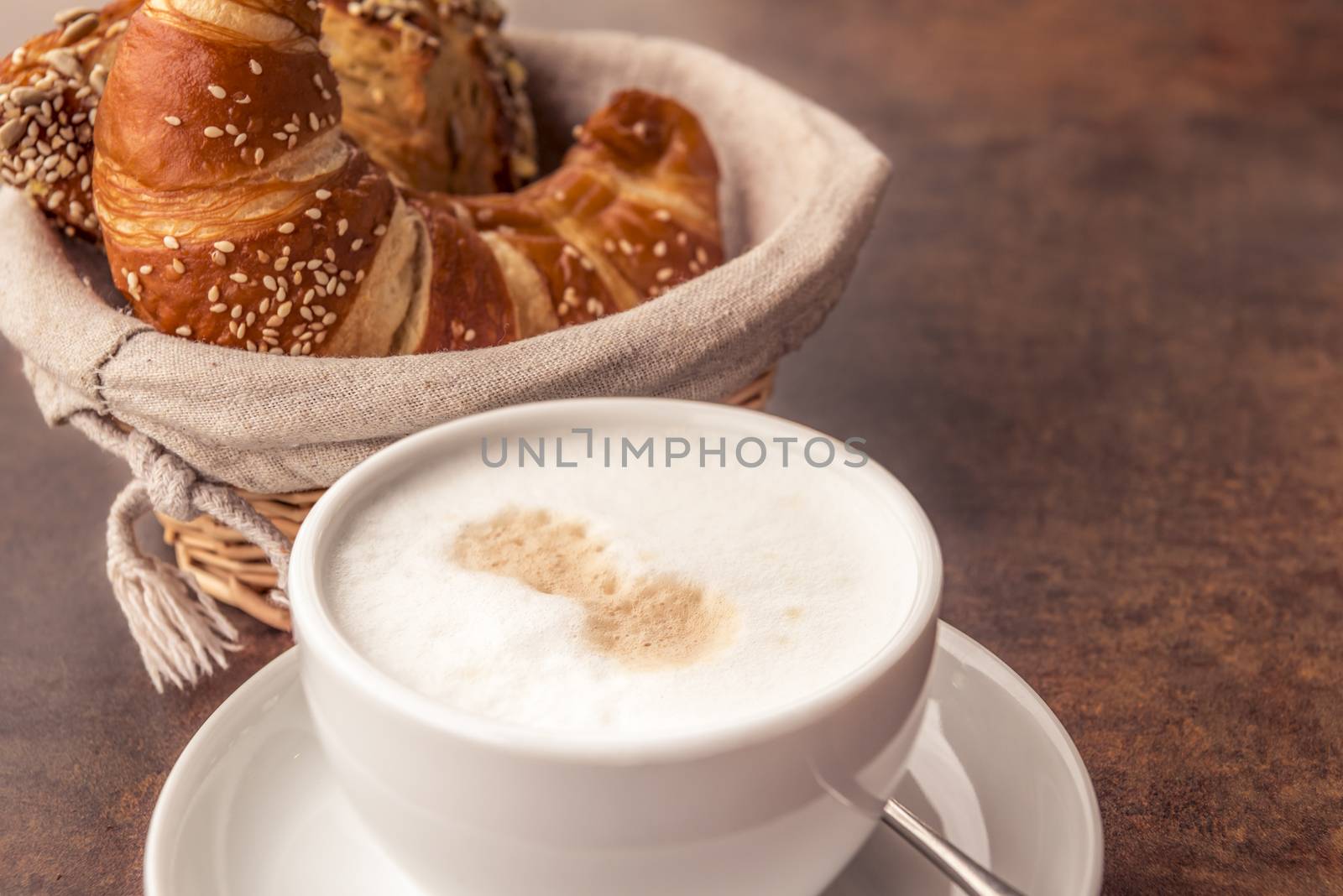 Delicious breakfast with a cup of milk coffee and fresh, sesame croissants in a rustic wicker basket, on a brown table.