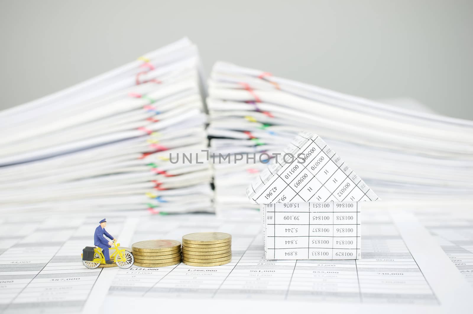 Postman is cycling pass step pile of gold coins and house on finance account have blur pile overload paperwork of report and receipt with colorful paperclip as background.