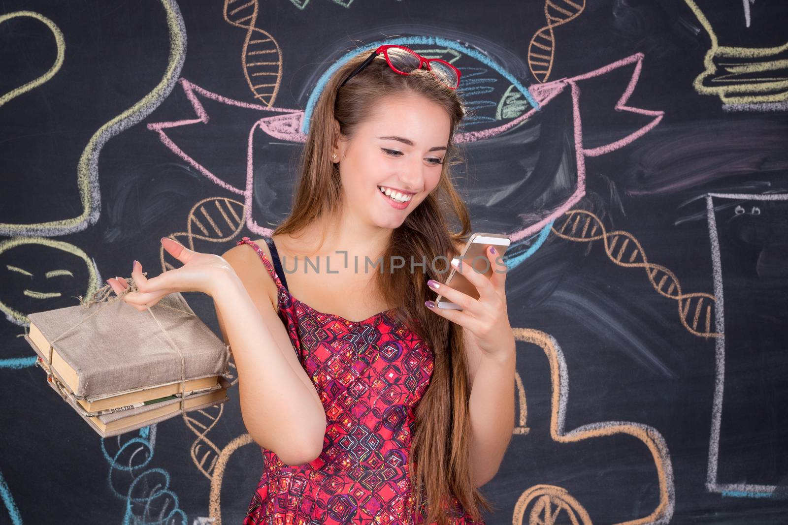 Young student girl in red dress with red glasses and pack of books poses in front of school classroom blackboard