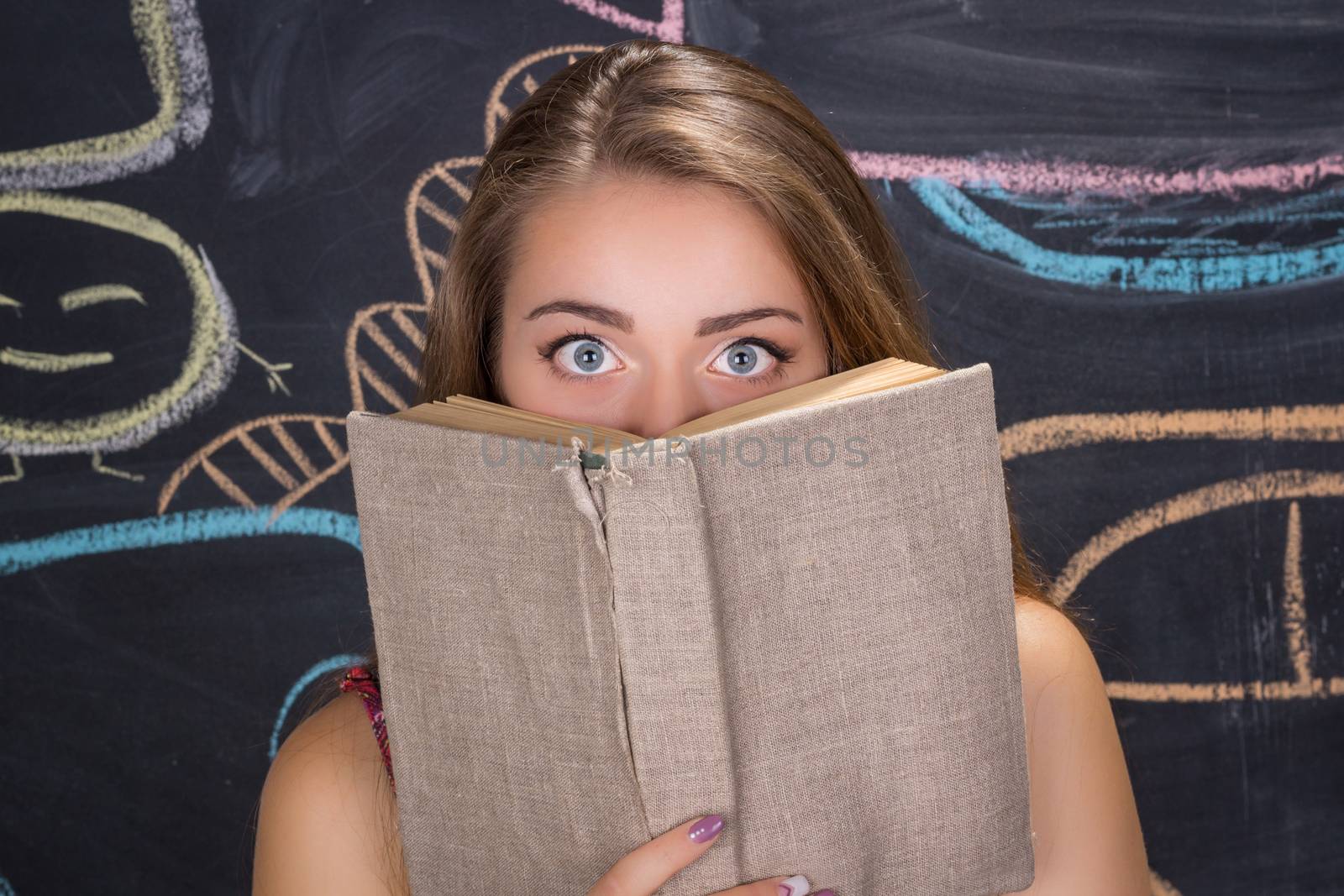 Perplexed  young student girl in red dress and red glasses reading a book hides behind it