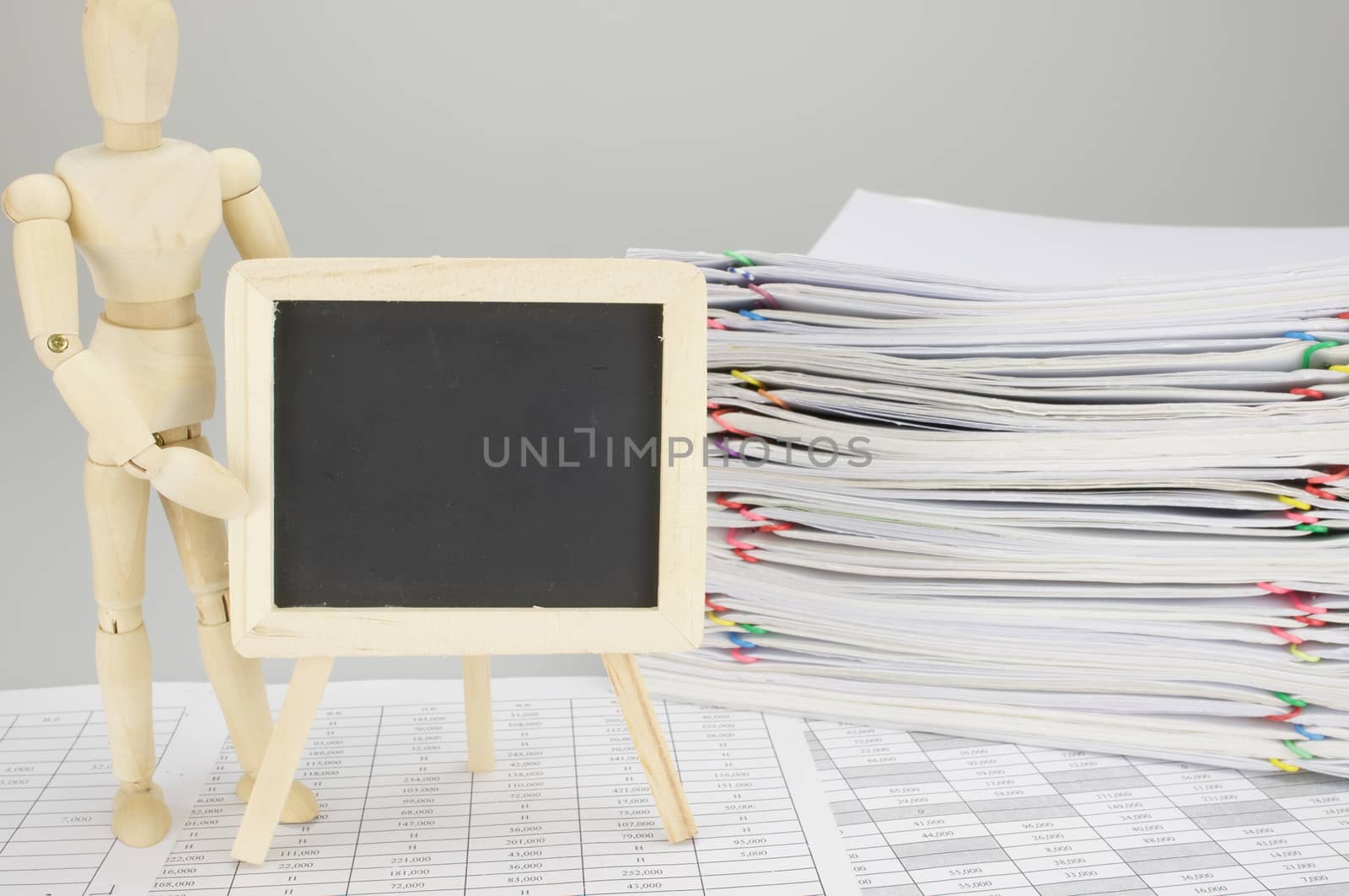 Wooden dummy holding empty blackboard on finance account have blur pile overload document of report and receipt with colorful paperclip as background.