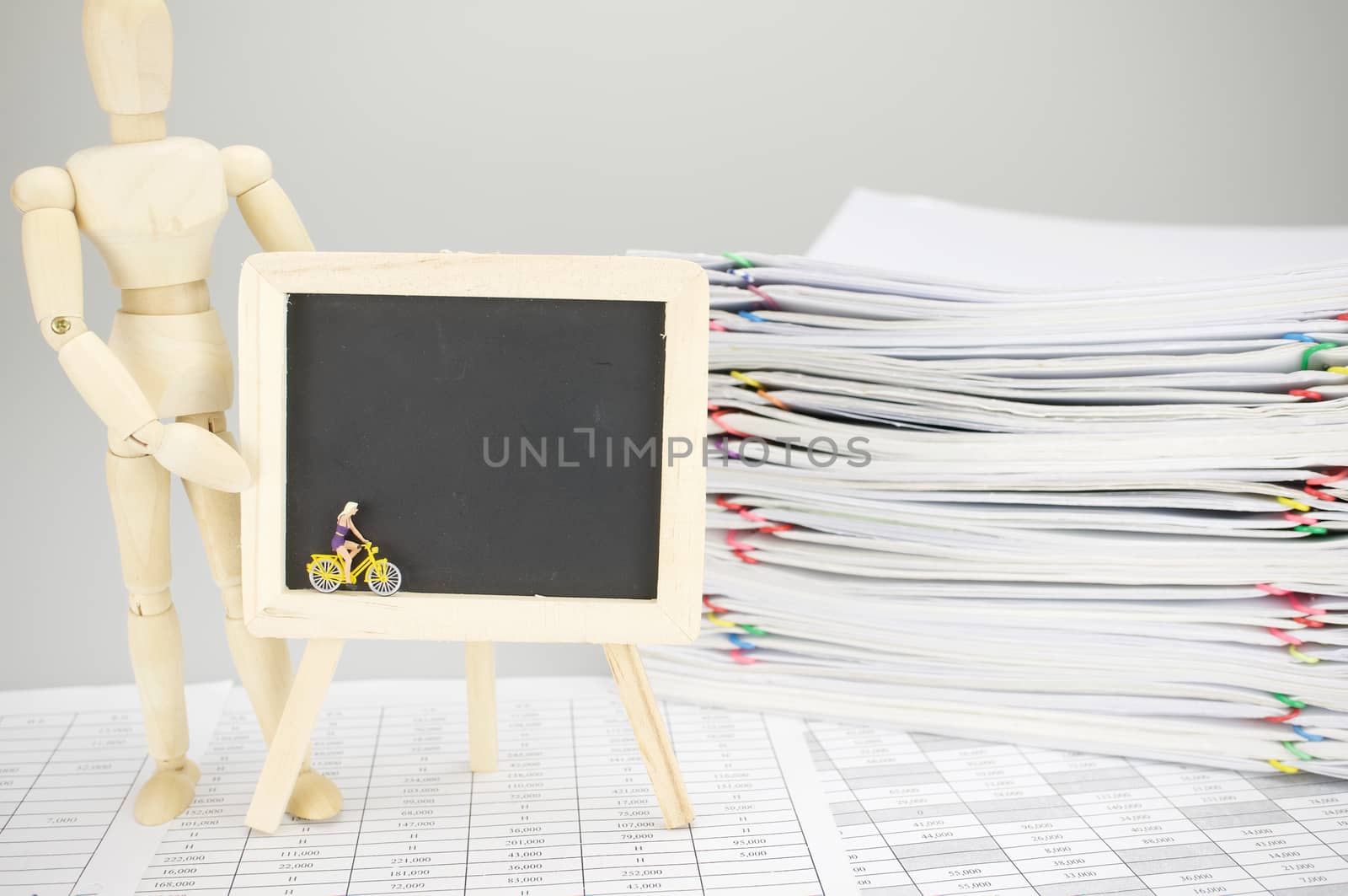 Wooden dummy holding empty blackboard with woman is cycling on finance account have blur pile overload document of report and receipt with colorful paperclip as background.